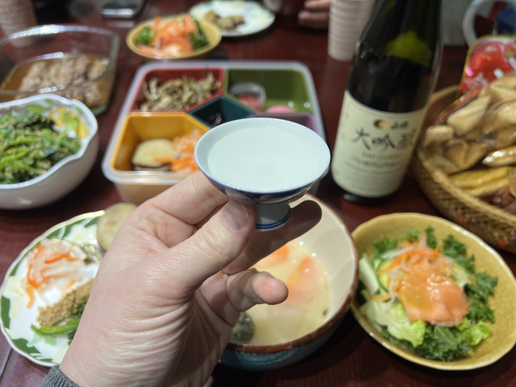 Chad holds up a saucer of sake over an assortment of colourful foods (with a Daiginjo sake bottle in the background)