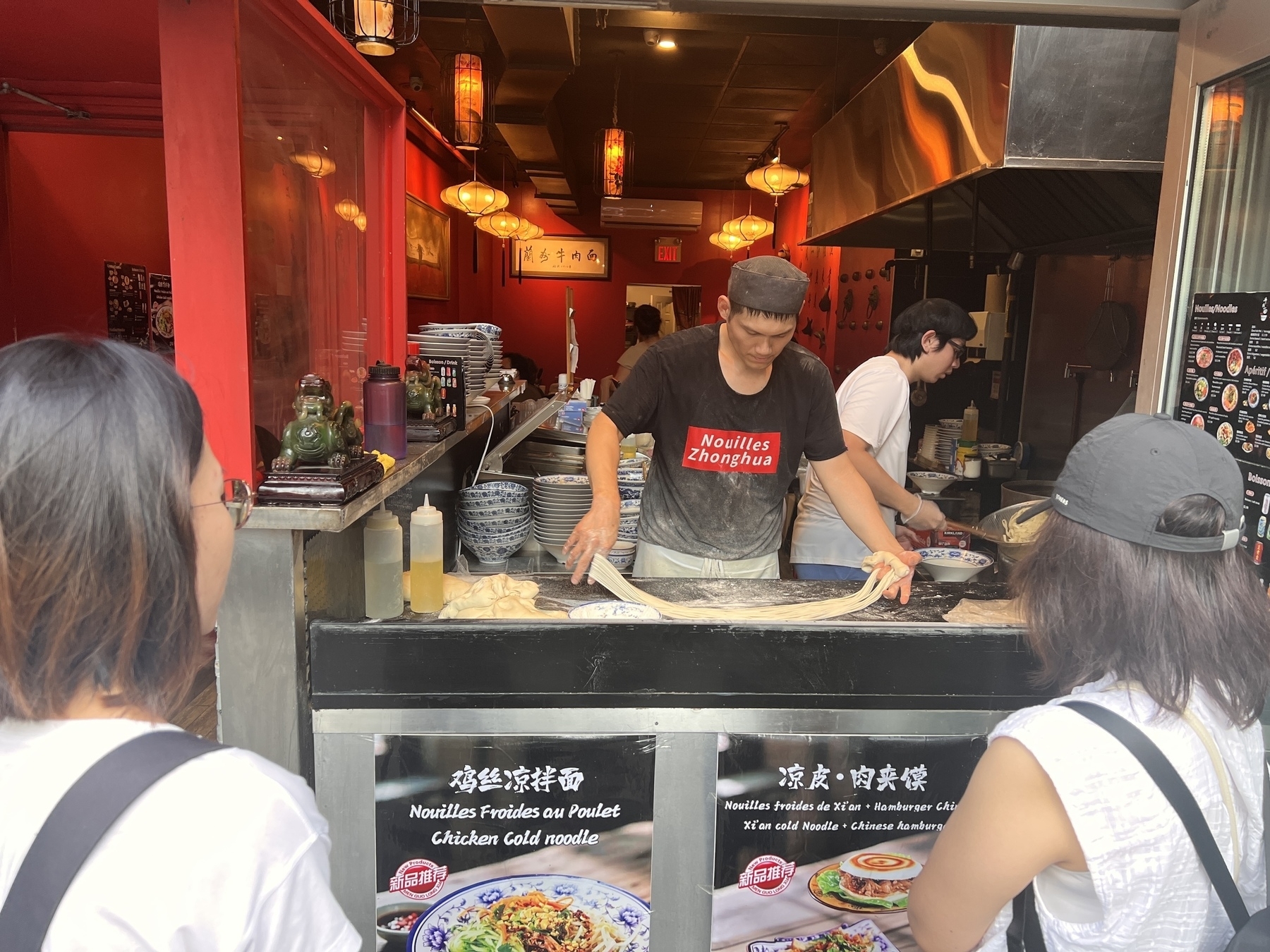 A man makes noodles at a Chinese noodle shop while two women look on. It’s like magic! 🪄 