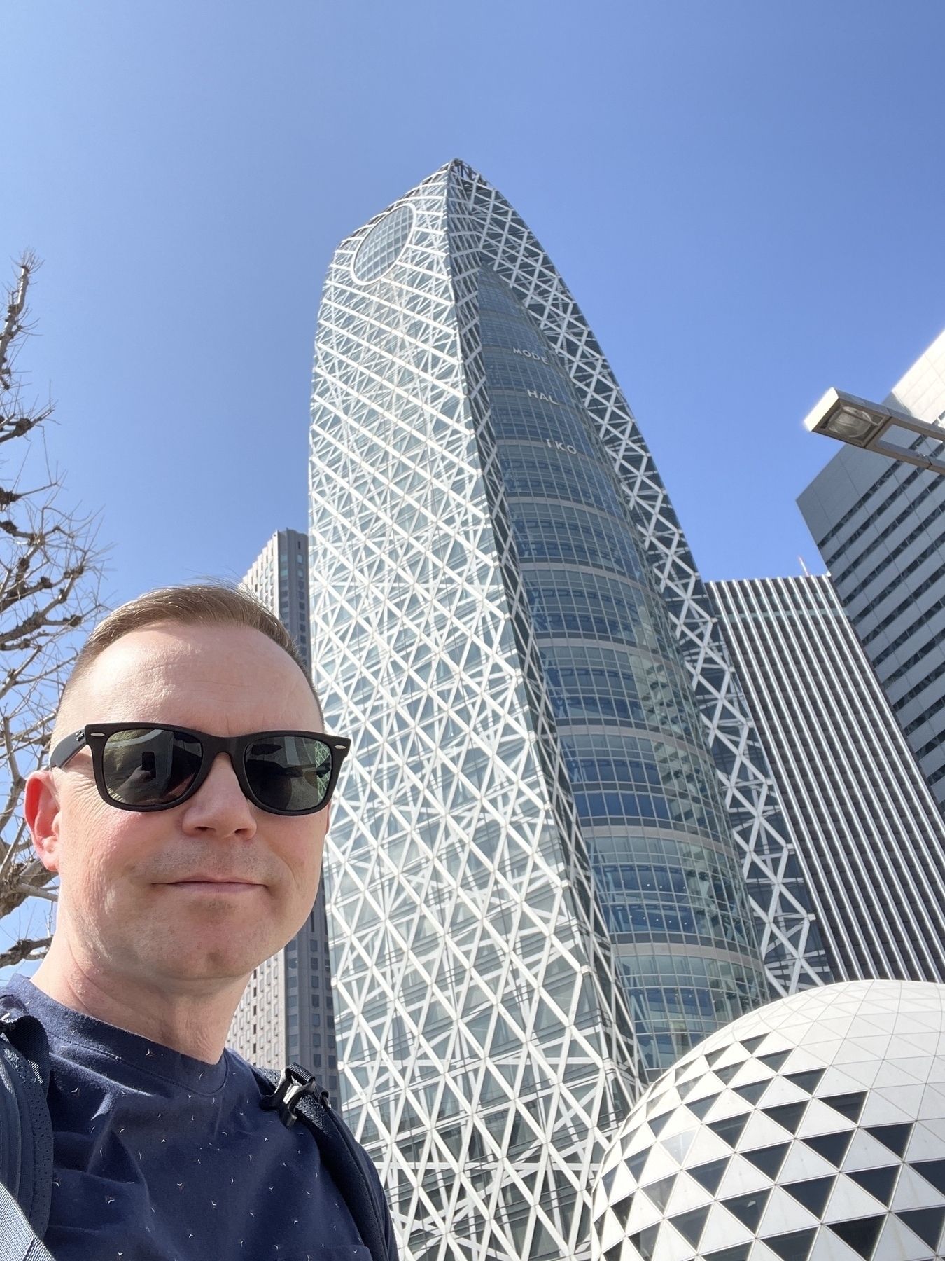 Chad selfie in front of the Mode Gakuen building against a clear blue sky. He is wearing sunglasses and a t-shirt. 