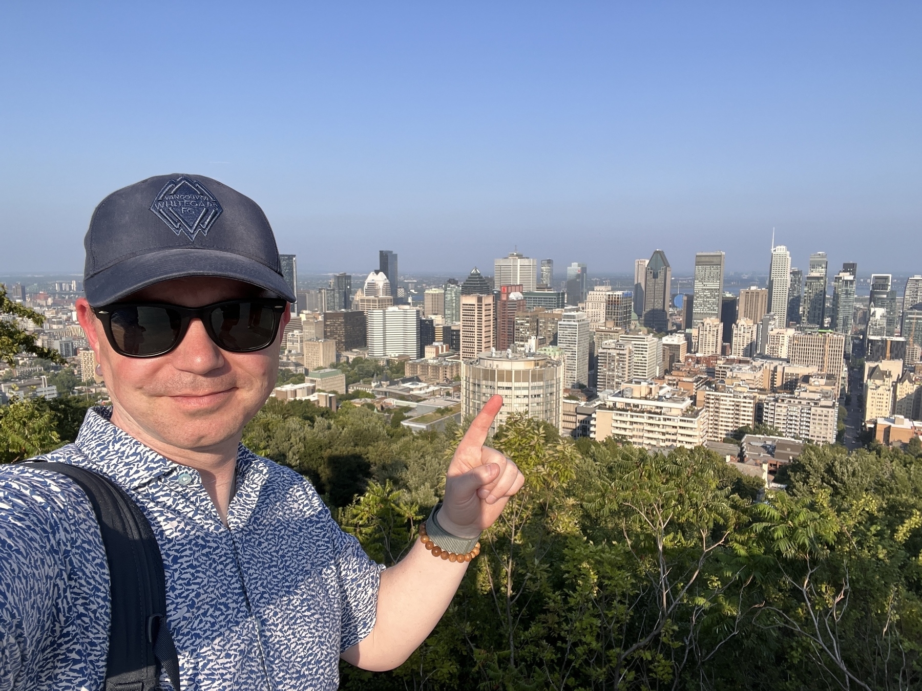 Chad selfie with a very red face pointing to downtown Montreal behind and below him. Cheekily he wears a Vancouver Whitecaps hat