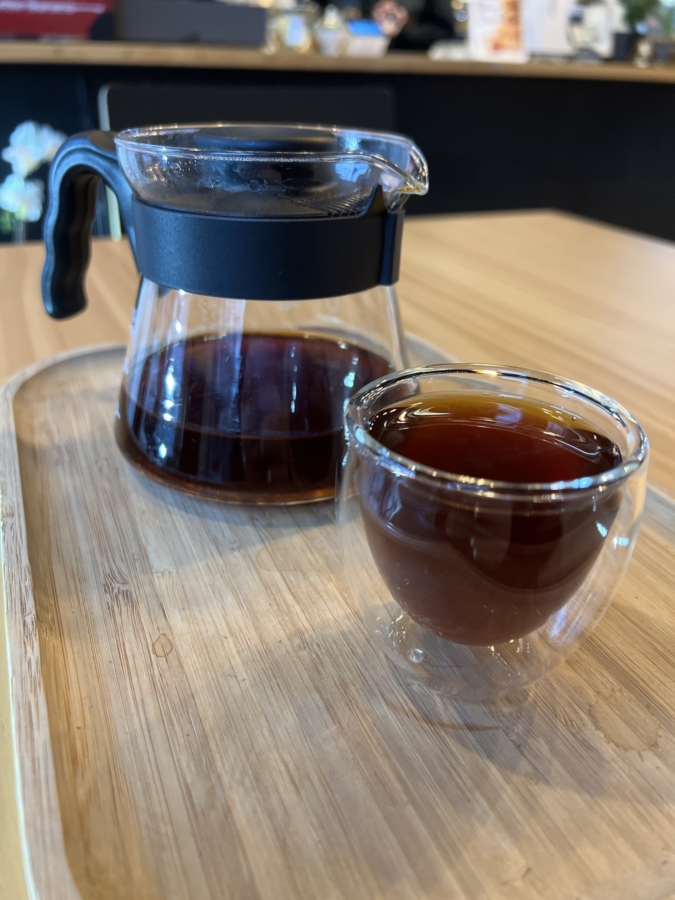 Small double walled glass tumbler with a glass Hario serving pot in the background, both vessels full of the clear brown liquid bean juice, sitting atop of a wooden tray. It is meditation time!