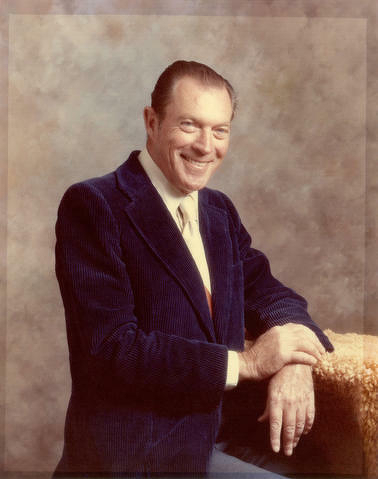 Gerald Sutton Brown portrait showing a white man smiling leaning on some sort of stand in a photo studio. The image is in colour