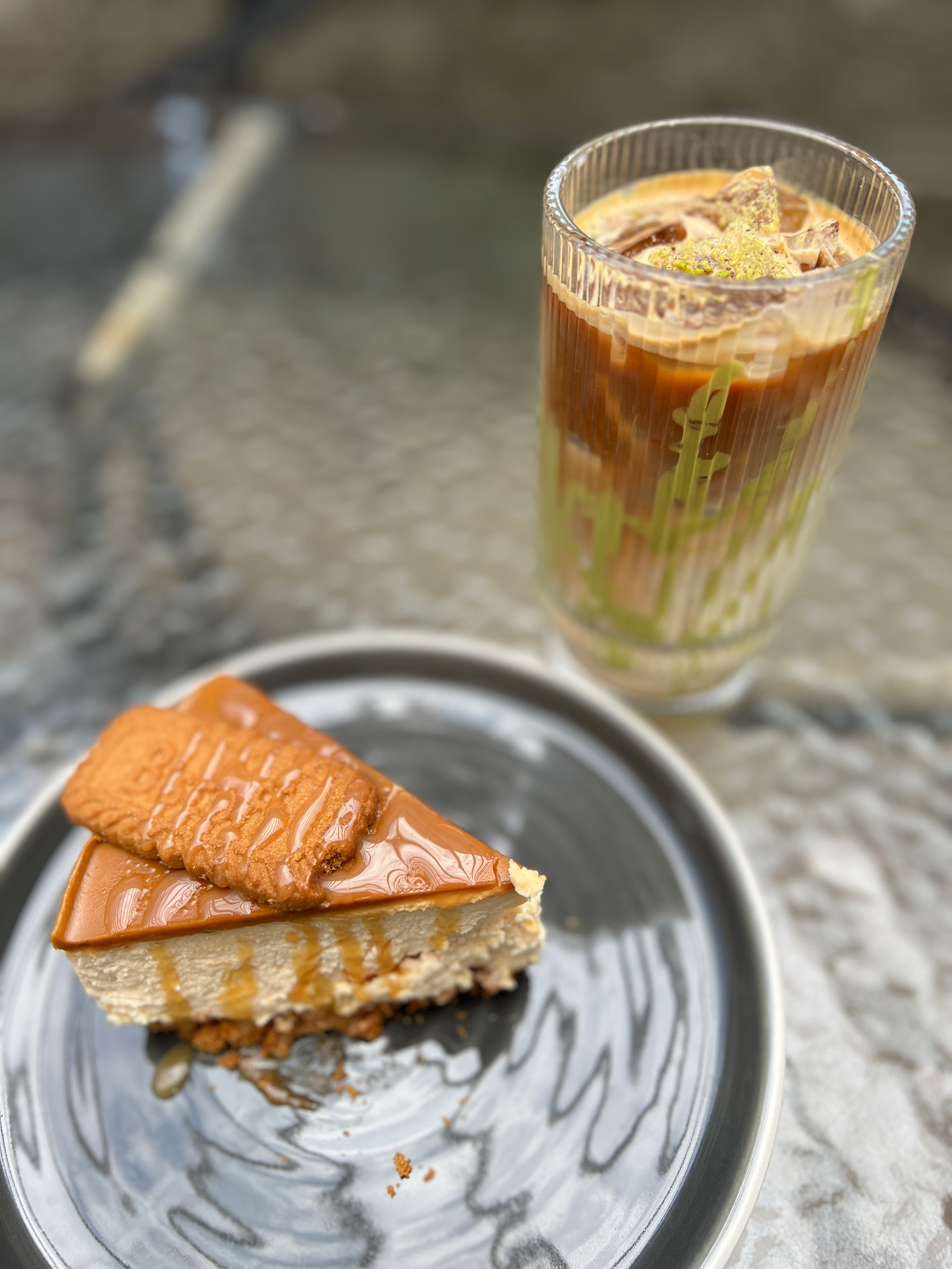 A slice of caramel-topped Biscoff cheesecake served on a gray plate alongside a layered iced pistachio and rose water latte in a tall, textured glass.