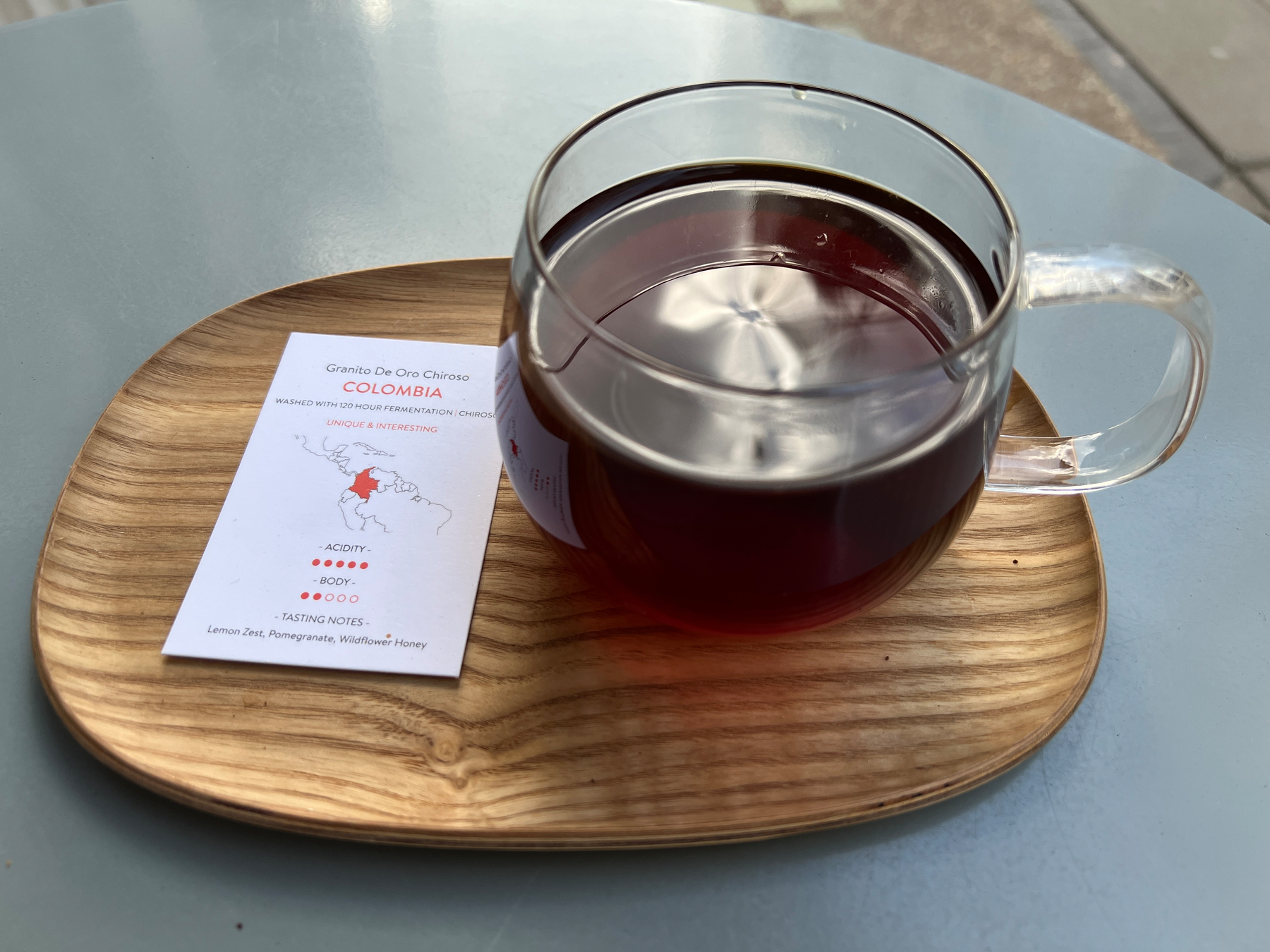 A glass mug full of coffee in a wooden tray with a business-card sized info sheet on the Granito De Oro Chirosso beans from Colombia
