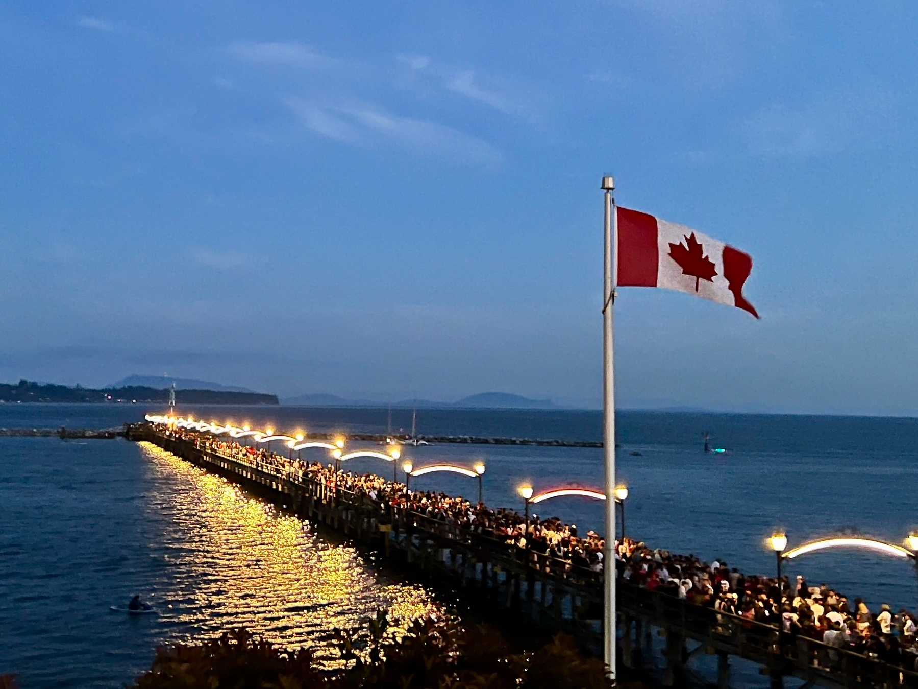 Canada flag flying over a lit up pier with tons of people on it