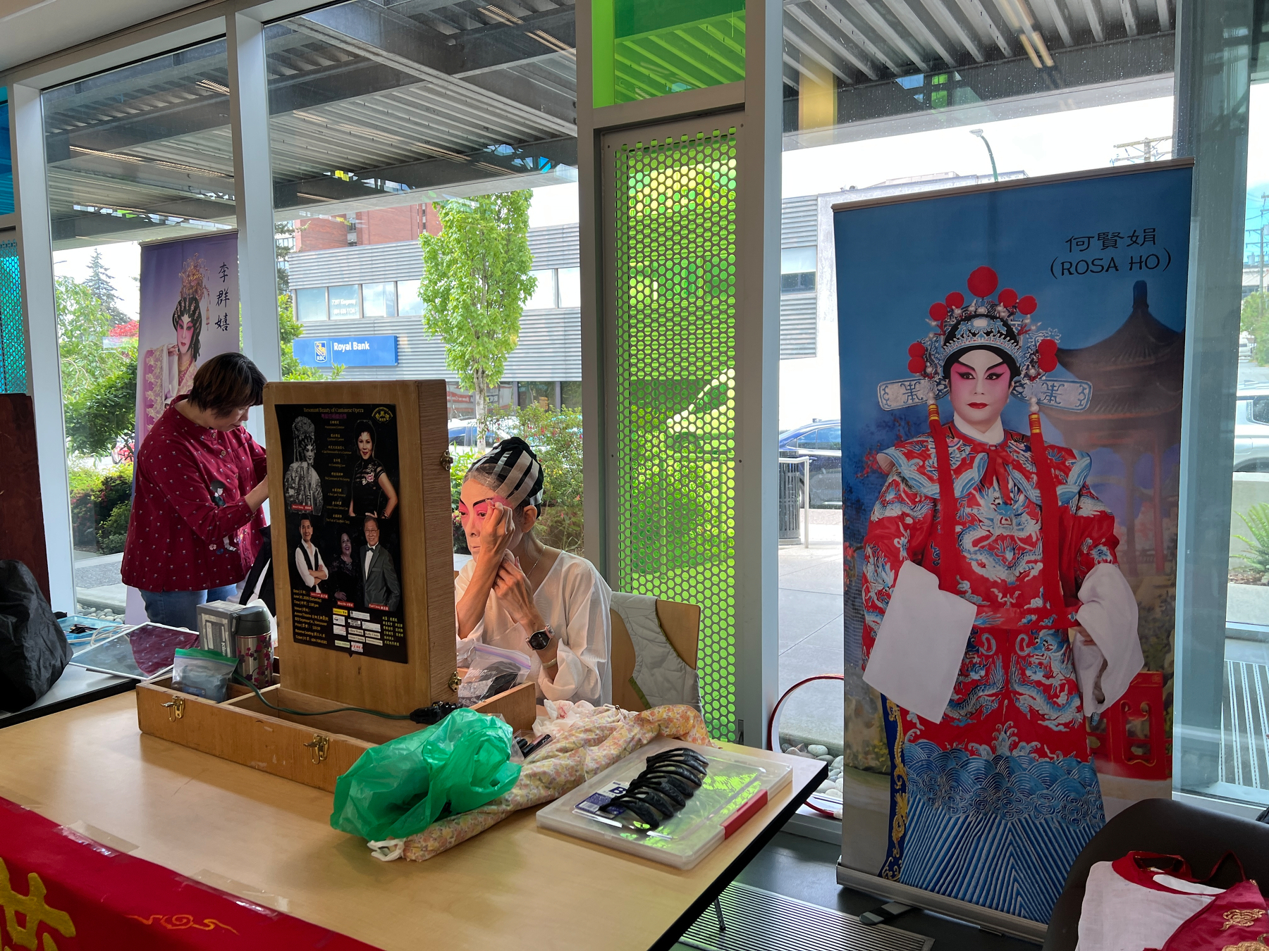 A performer puts on makeup. Beside her is a banner poster of an opera character in full regalia
