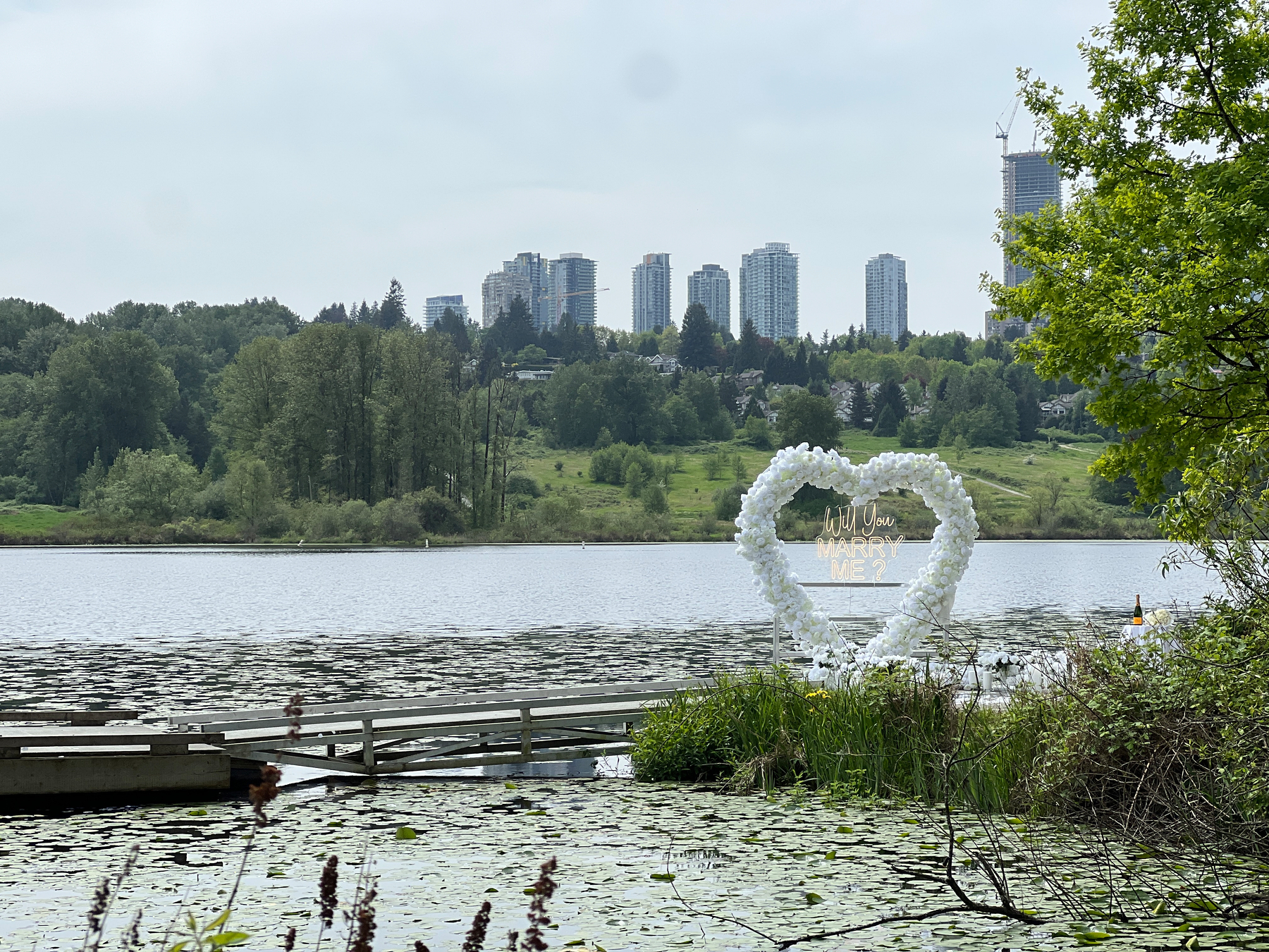 In a dock on the lake is a large heart made of flowers with a lit up sign “Will you marry me?”&10;&10;In the background across the lake are the condo towers of Metrotown