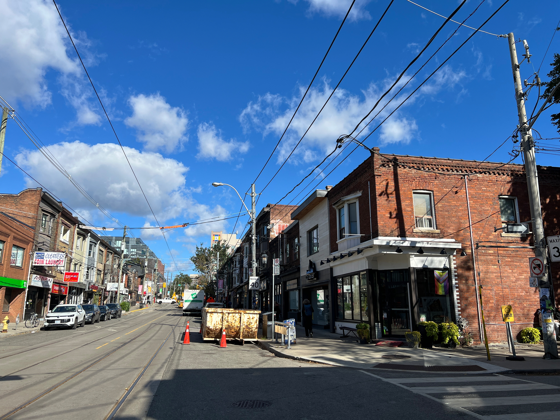 A sunny urban street scene features brick buildings, parked cars, overhead utility lines, and a clear blue sky with scattered clouds.