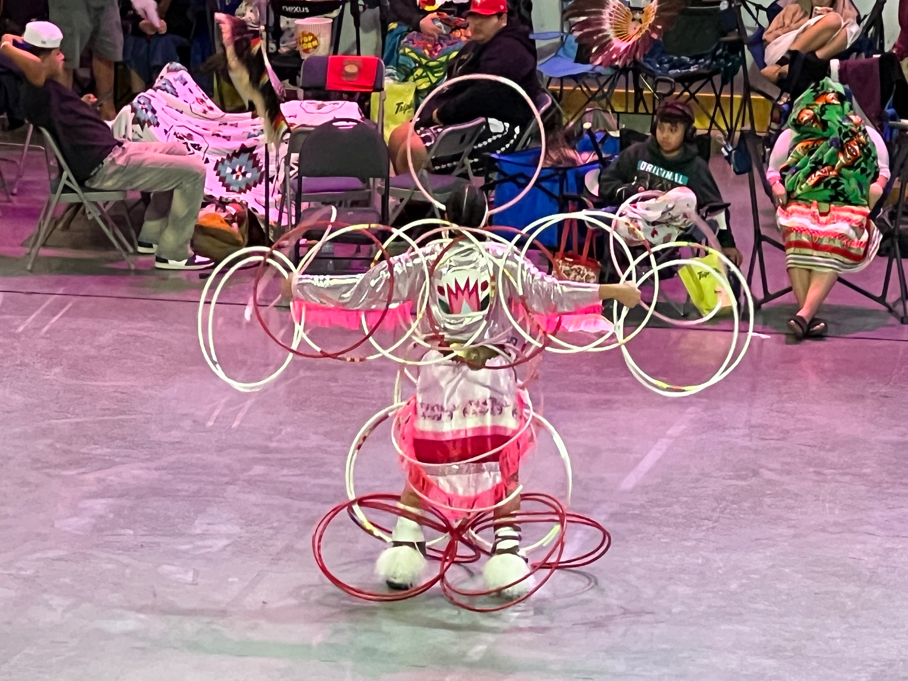 A performer dressed in traditional attire is skillfully manipulating multiple hoops simultaneously in front of an audience seated on chairs.