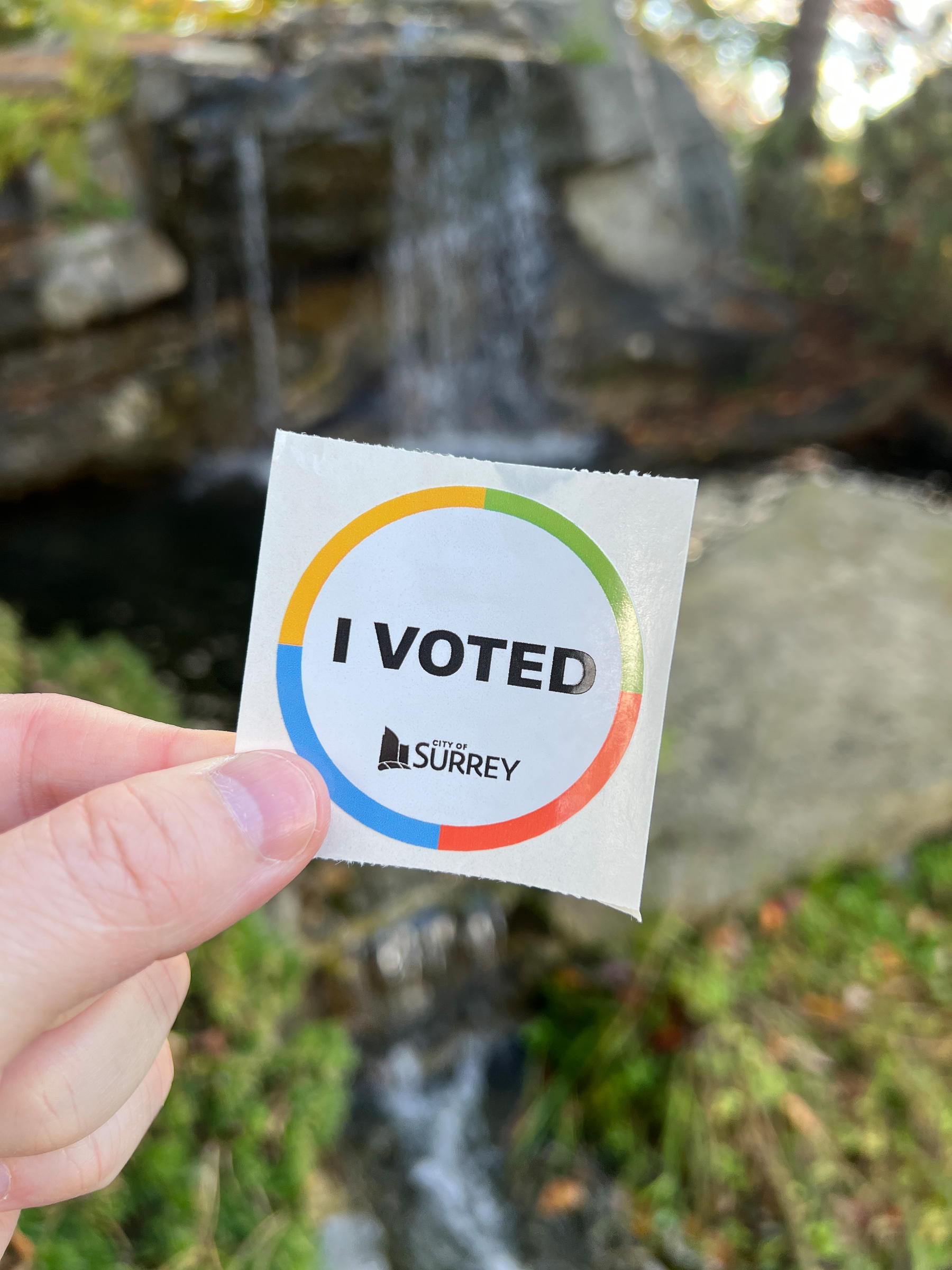 A hand is holding an I Voted sticker from the City of Surrey with a blurred waterfall and natural surroundings in the background.