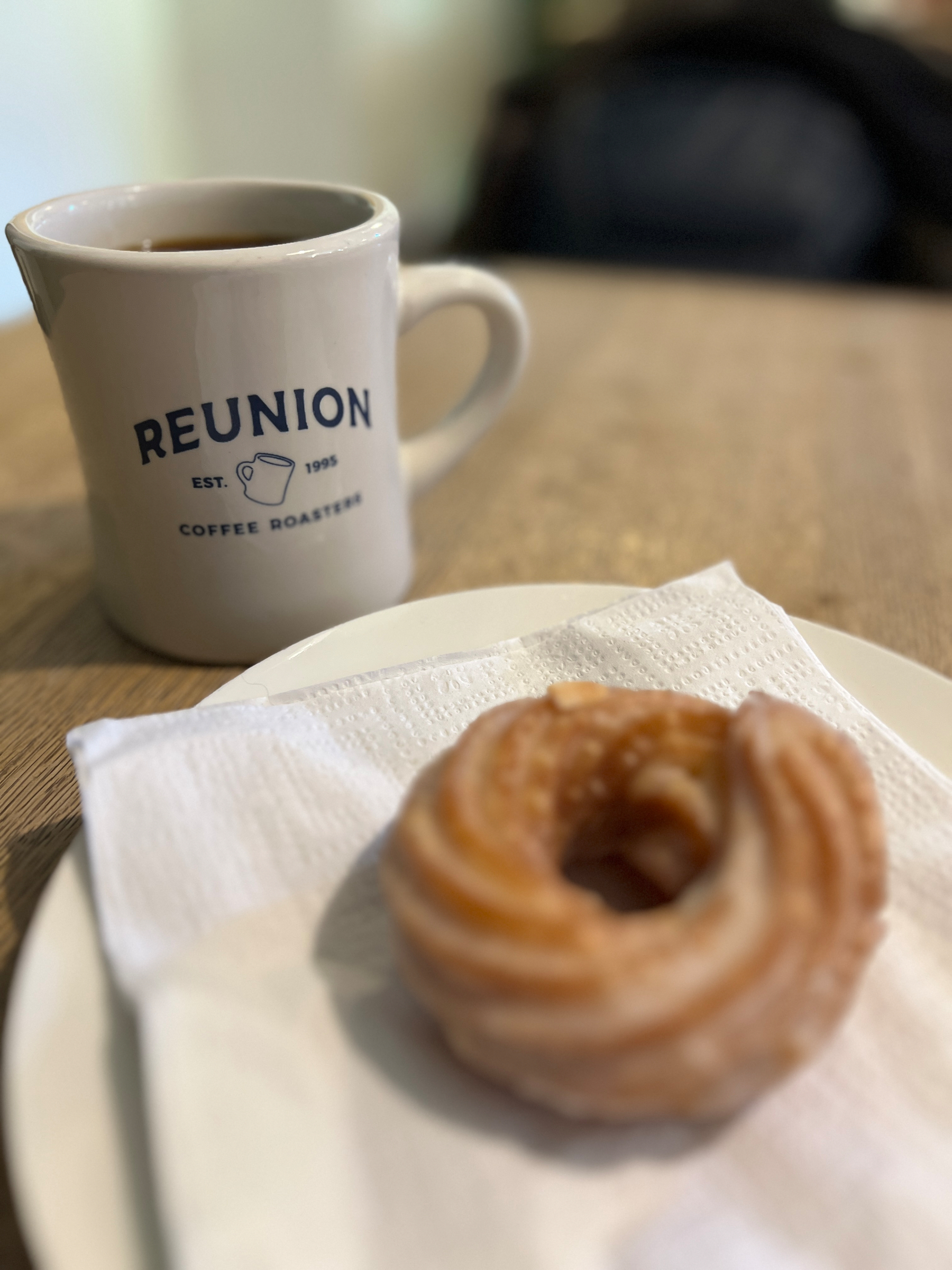 A mug of coffee from Reunion Roasters beside a glazed donut resting on a white napkin on a plate atop a wooden table.