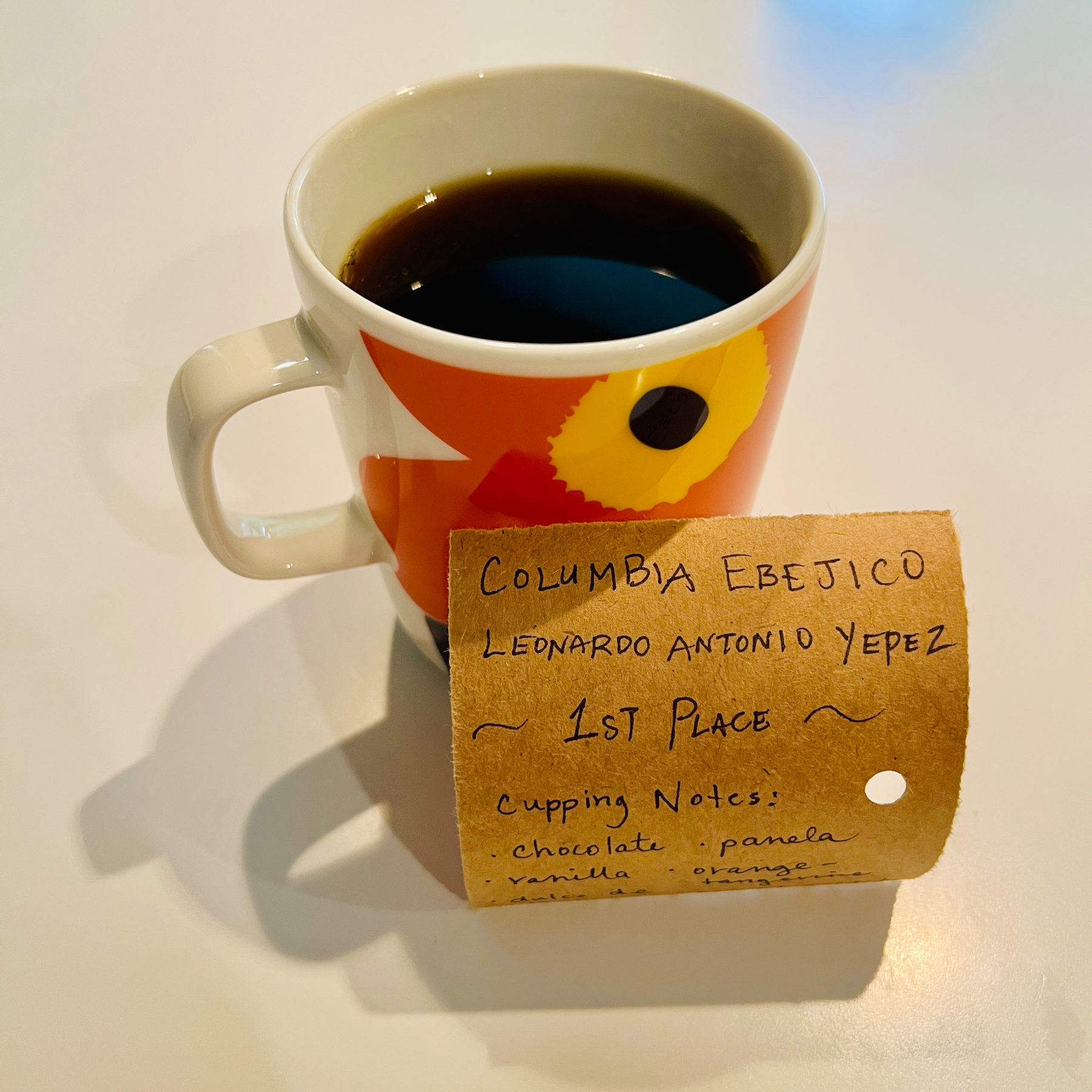 A cup of coffee with a handwritten label leaning against it: COLUMBIA EBEJICO LEONARDO ANTONIO YEPEZ ~ 1ST PLACE ~