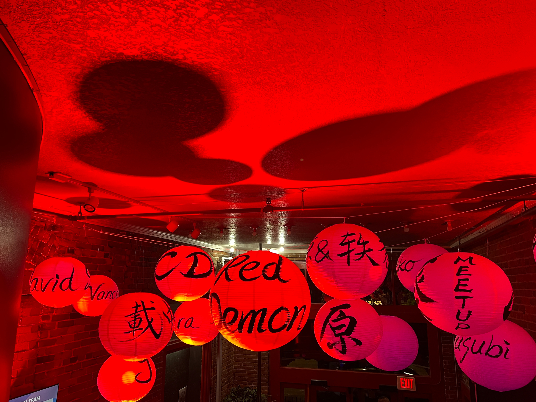 Red lit paper lanterns with various English and Japanese characters hang from the ceiling, casting shadows on a red-lit room. The middle one says Red Demon