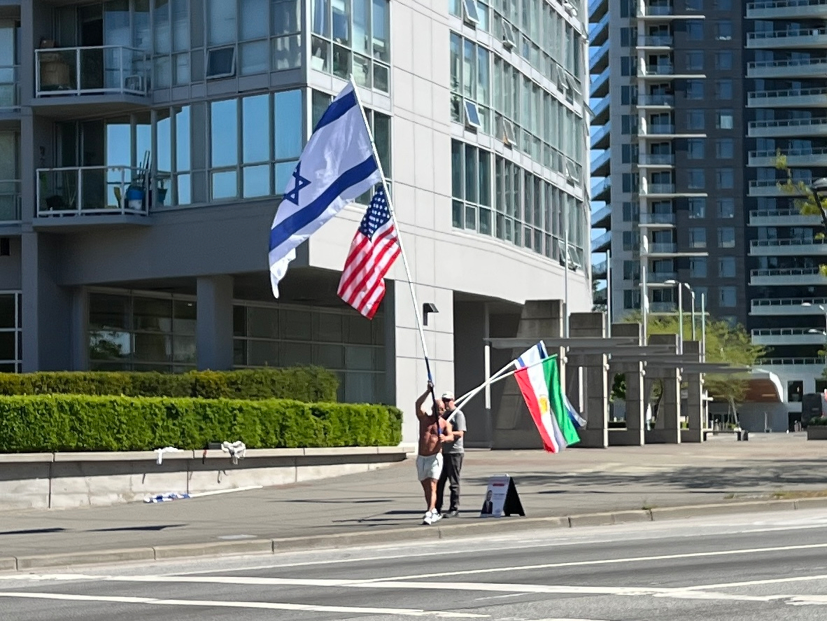 Two guys one holding a tall flagpole with the Israel and US flags, another holding two flags: Israel and the Iran flag with the monarchist lion symbol
