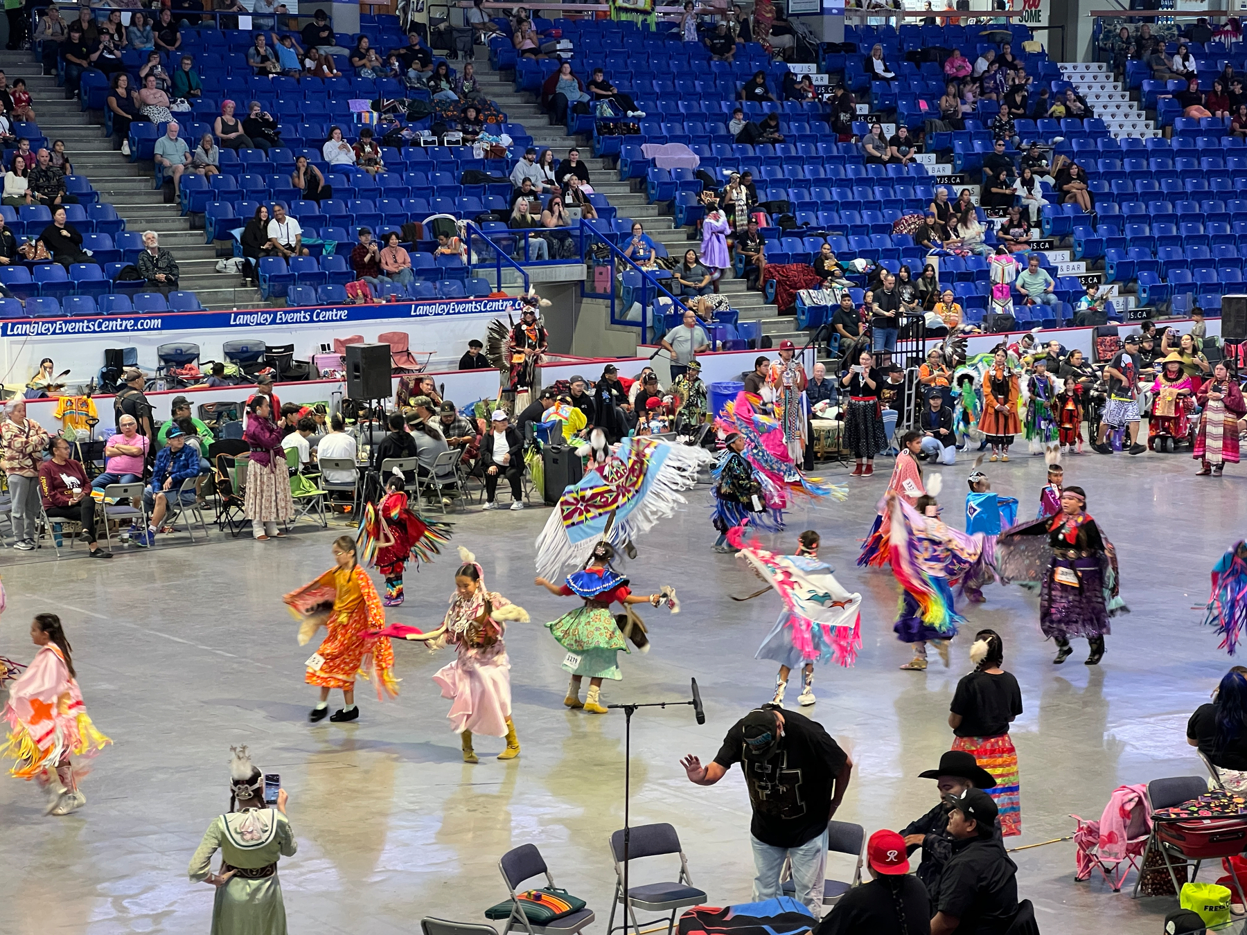 A group of kids dressed in colorful traditional regalia are dancing in a large indoor arena with spectators seated in the stands and along the perimeter.