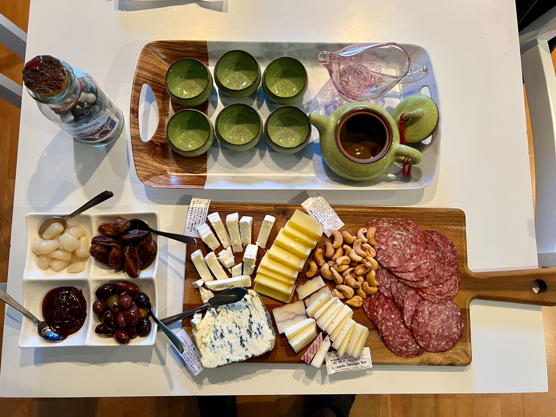 Charcuterie board with bison salami, cashews, 4 kinds of cheese. A plate with pickled onions, dates, olives, and spicy cranberry jam. A tray with Chinese tea set. A jar full of candies from Heidelberg
