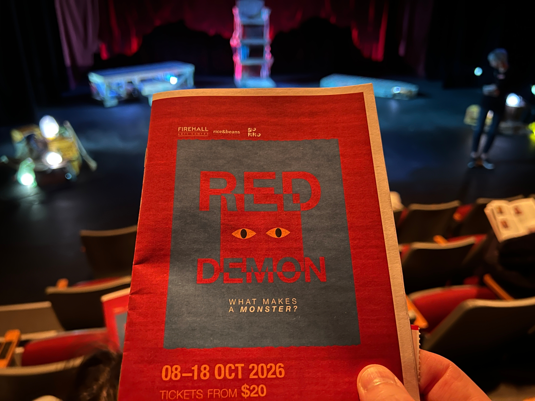 A hand is holding a red and gray program for a production titled Red Demon with the tagline What makes a monster? inside a theater with a stage and seating area in the background.