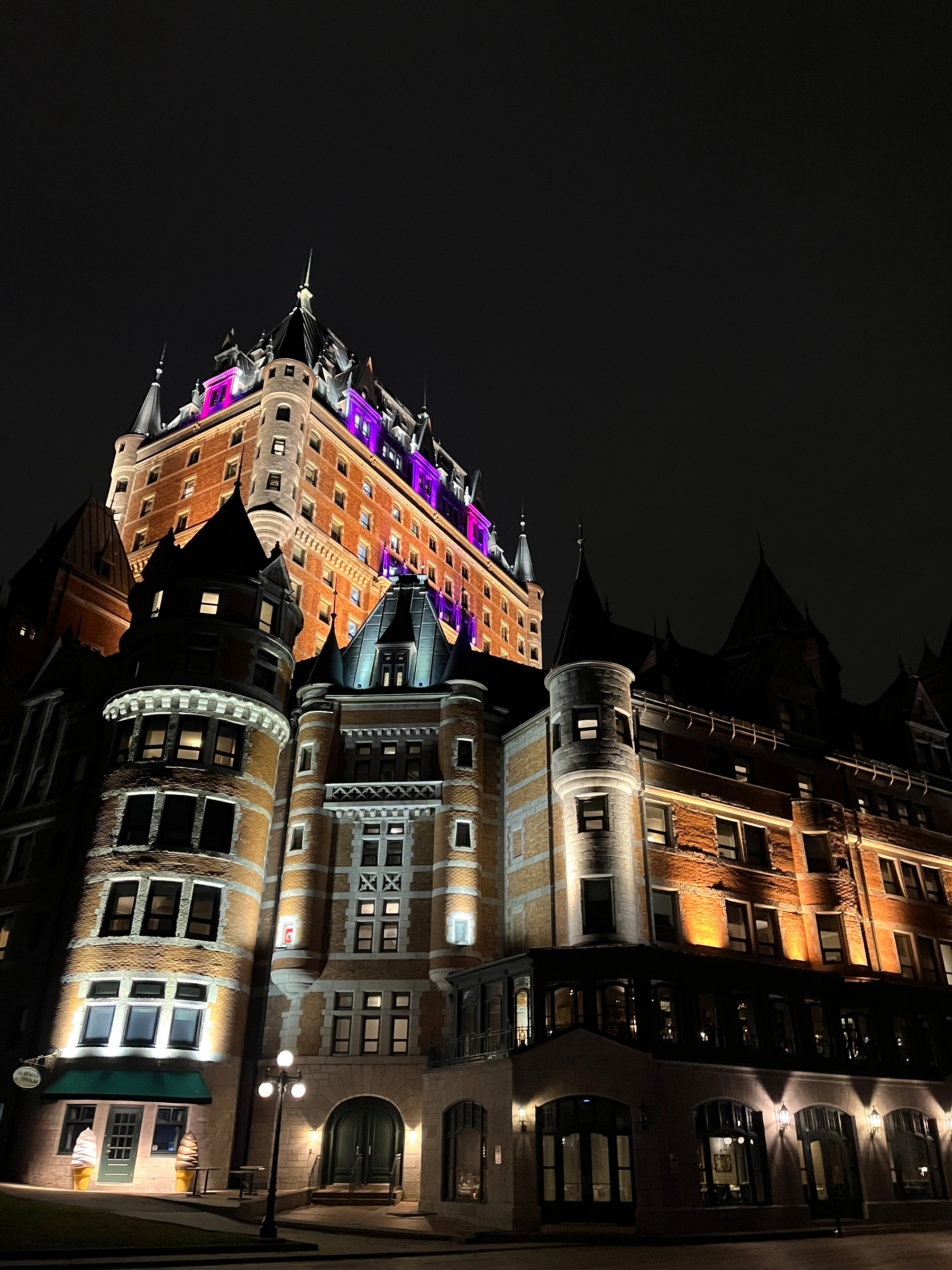 A grand, historic building with turrets and illuminated facades stands against a dark night sky, with some upper windows lit in purple.