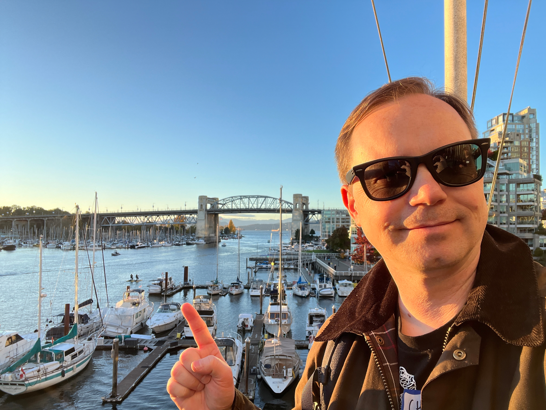 Chad wearing sunglasses is smiling and pointing towards the marina filled with boats and Burrard Bridge in the background during a sunny day.