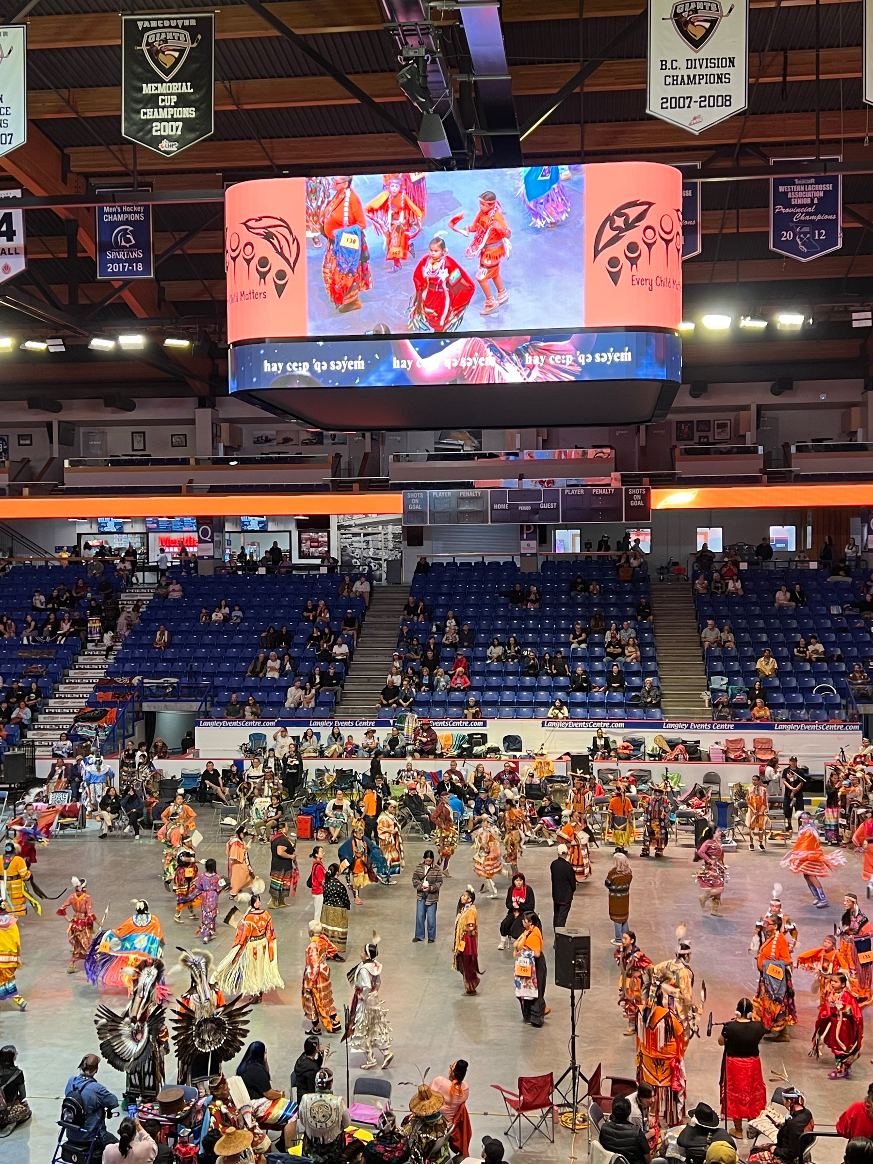 A large group of people dressed in colorful traditional Indigenous attire are participating in a powwow inside an arena with spectators seated in the stands.