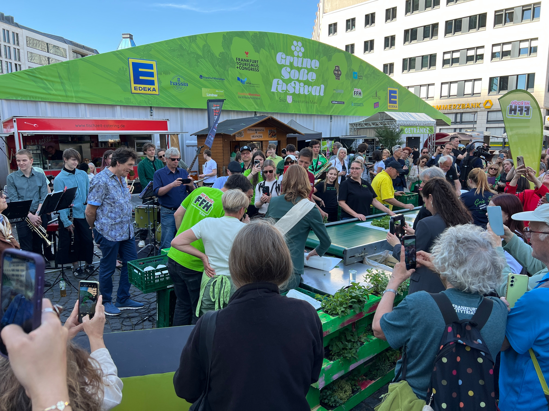People crowded around a little conveyor belt taking photos. In the back is a big sign for the Frankfurter Grüne Soße Festival