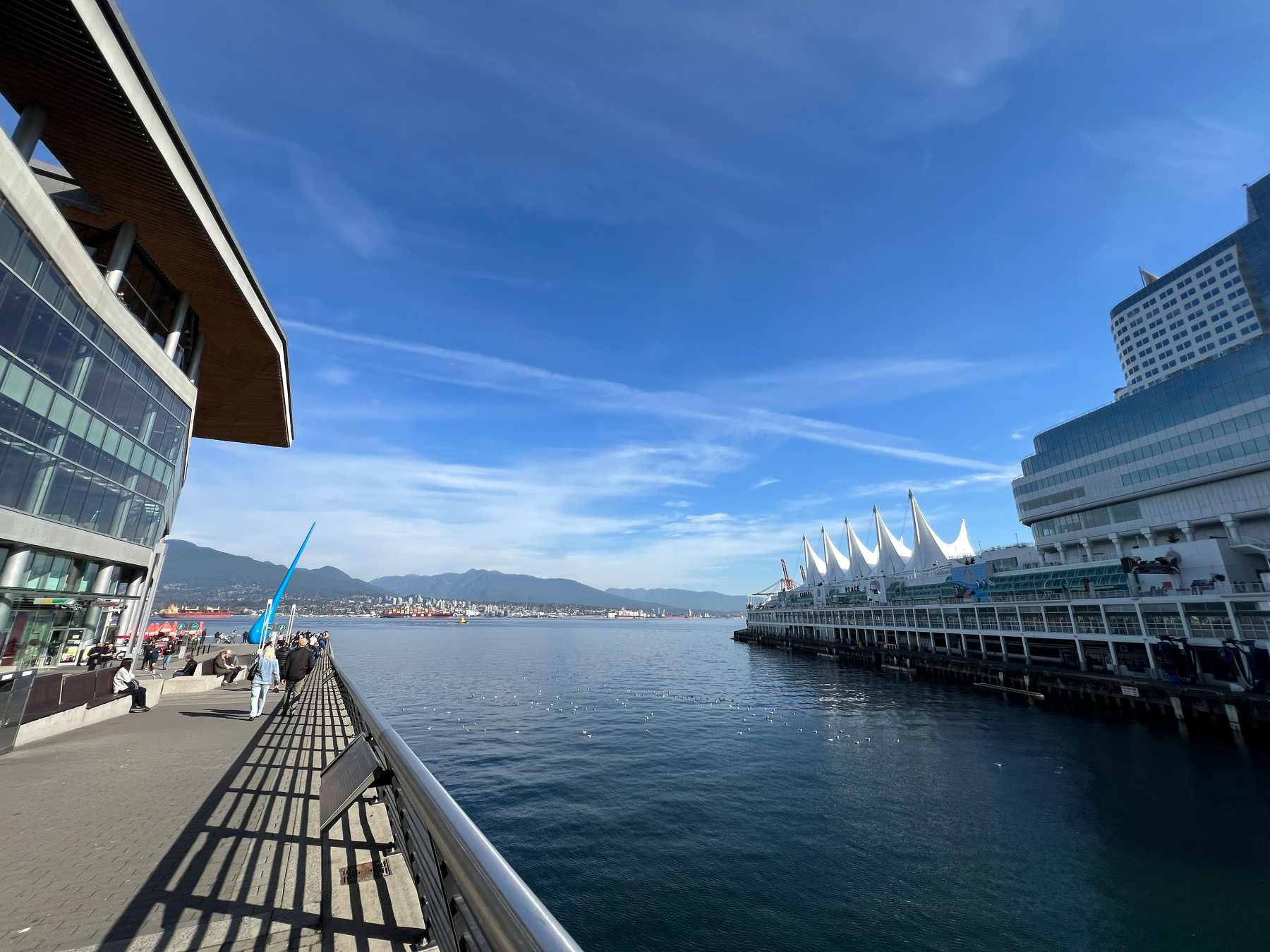 A waterfront scene featuring modern buildings, a boardwalk with people, calm water, and a clear blue sky with distant mountains.