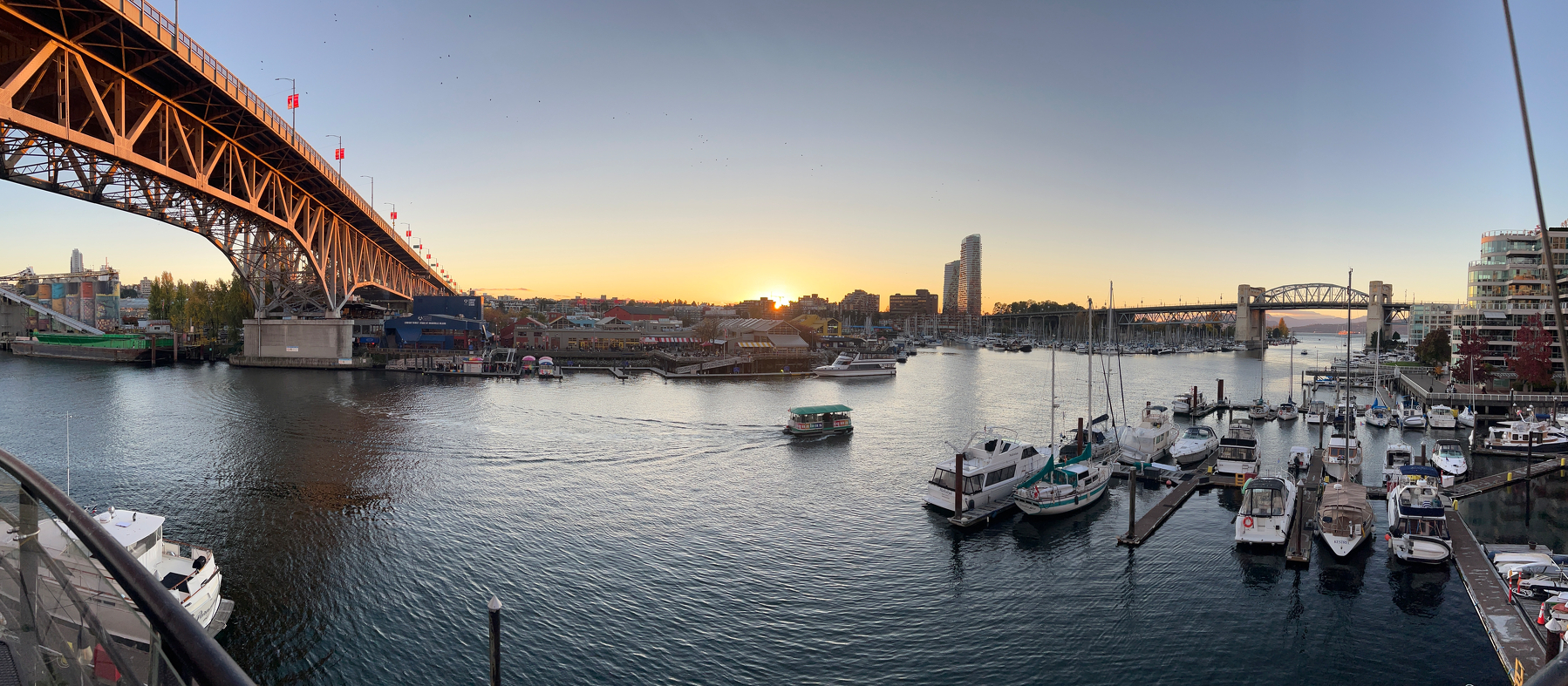 Pano of waterfront scene at sunset features a Granville Bridge on the left, boats docked at a marina on the right, and a Burrard Bridge in the distance.
