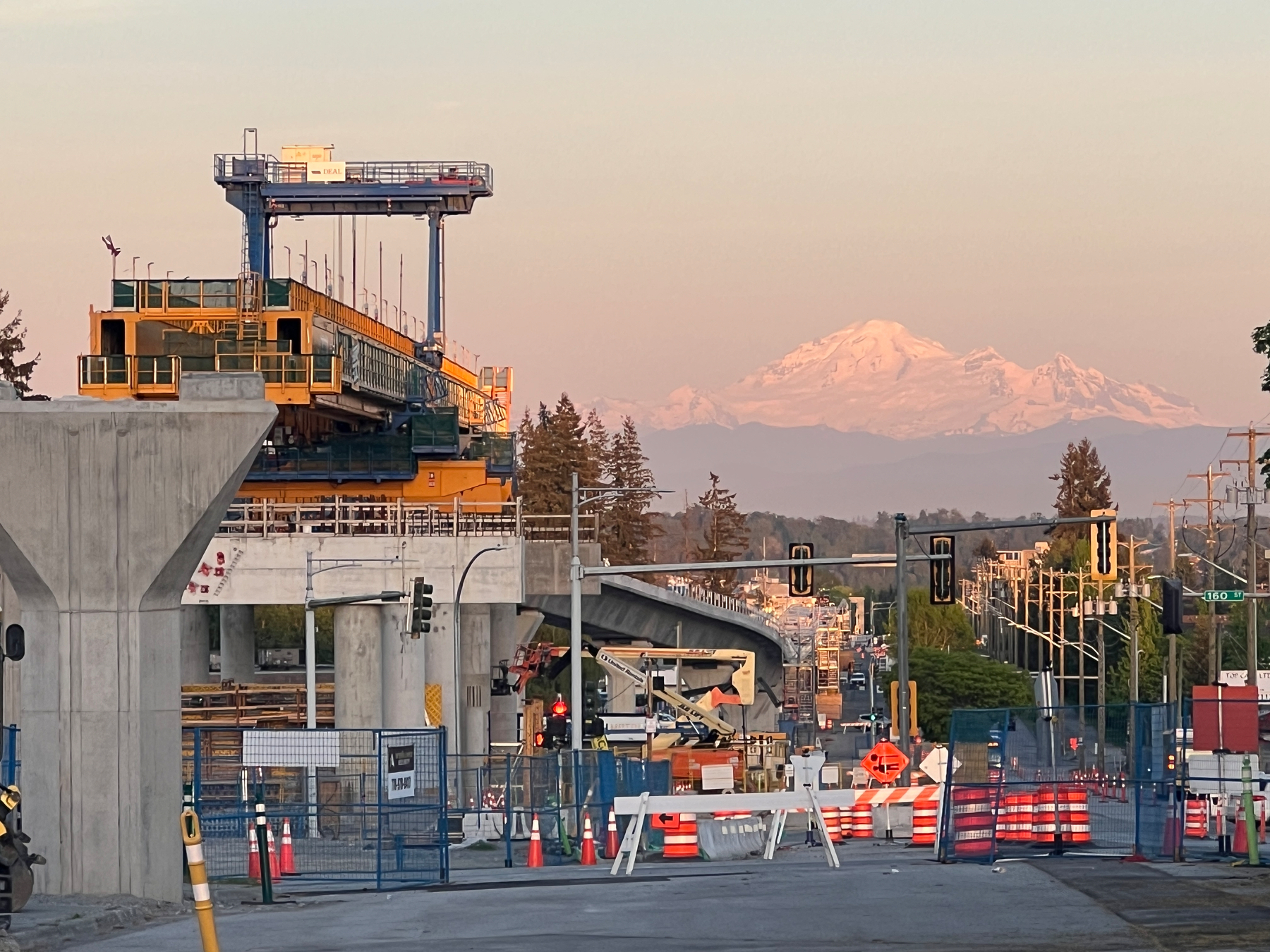 A launching gantry on the elevated SkyTrain track above a construction area. In the far distance is a large snowy mountain turning pink in the sunset