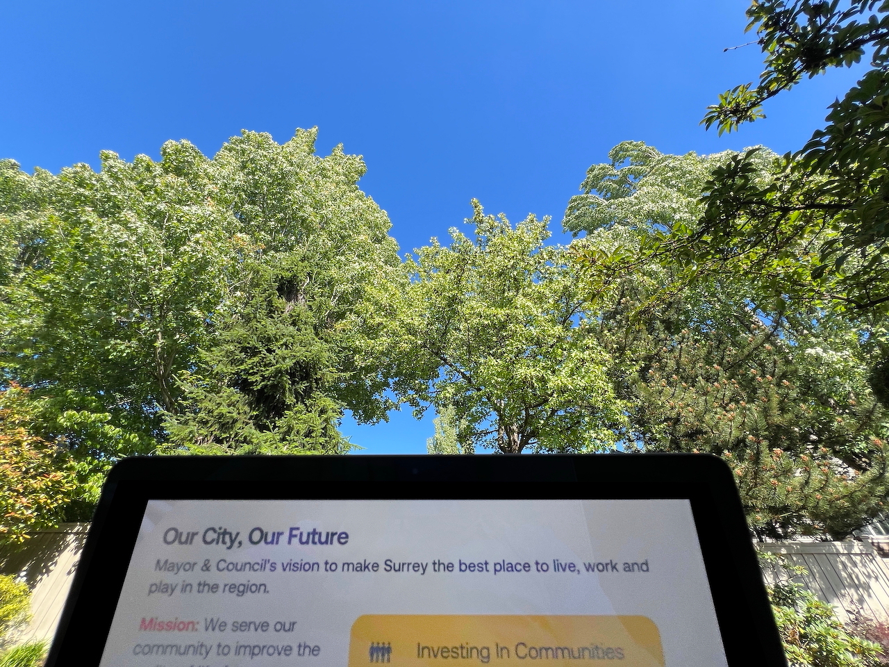 Wide angle of blue sky and trees. At the bottom of the frame is a computer with a slide titled: Our City, Our Future. Mayor & Council's vision to make Surrey the best place to live, work and play in the region.