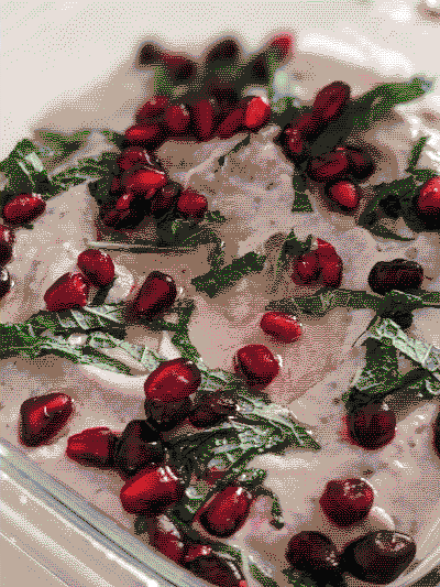 Closeup of mutabal in a square glass dish. The mutabal is a creamy dip in a very light brown/white color. It is topped with olive oil, chopped mint, and pomegranate seeds.