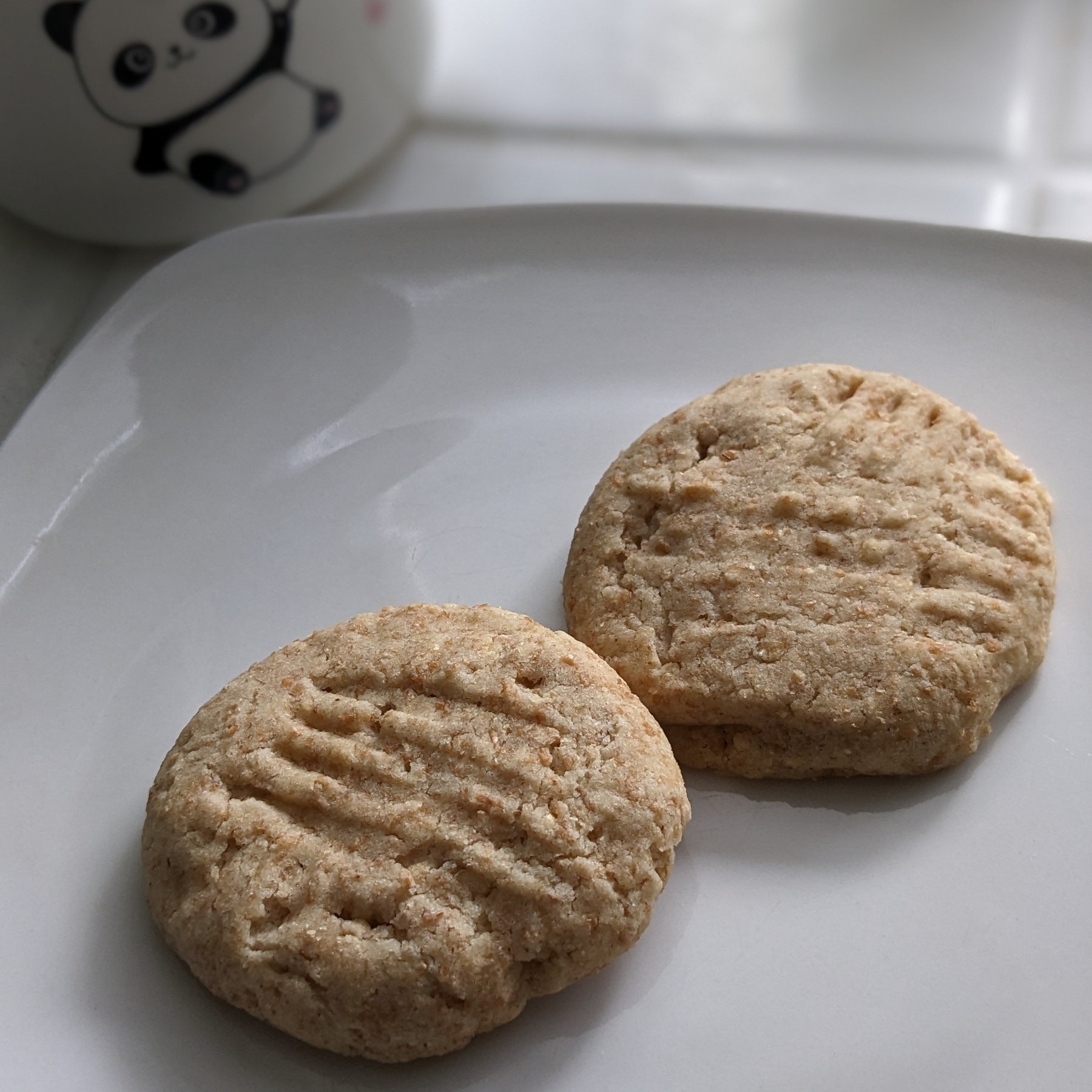 A photo of two light brown cookies sitting on a white plate. The cookies are homemade and have markings on top to show that they were pressed down with a fork. There is a white mug in the background with a cartoon panda on it.