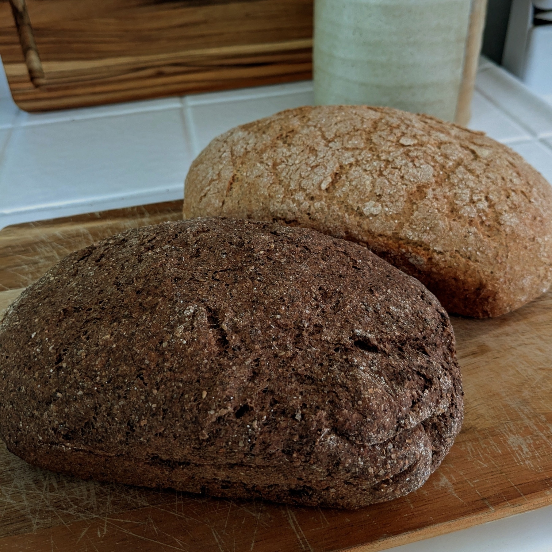 Two homemade loaves of bread sit on a wooden cutting board on a white tile kitchen counter. They are angled diagonally across the frame. The loaf in the front is a darker brown while the second loaf is much lighter in color. 