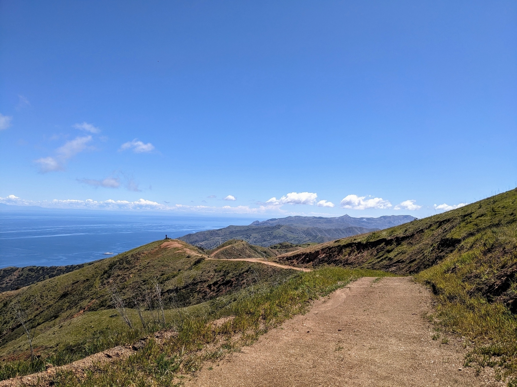 Photo of a hiking path that winds away into the distance. It is high elevation and can be seen following the tops of hills. The ocean can be seen on the far left corner and the sky above with clouds low and close to hills.&10;