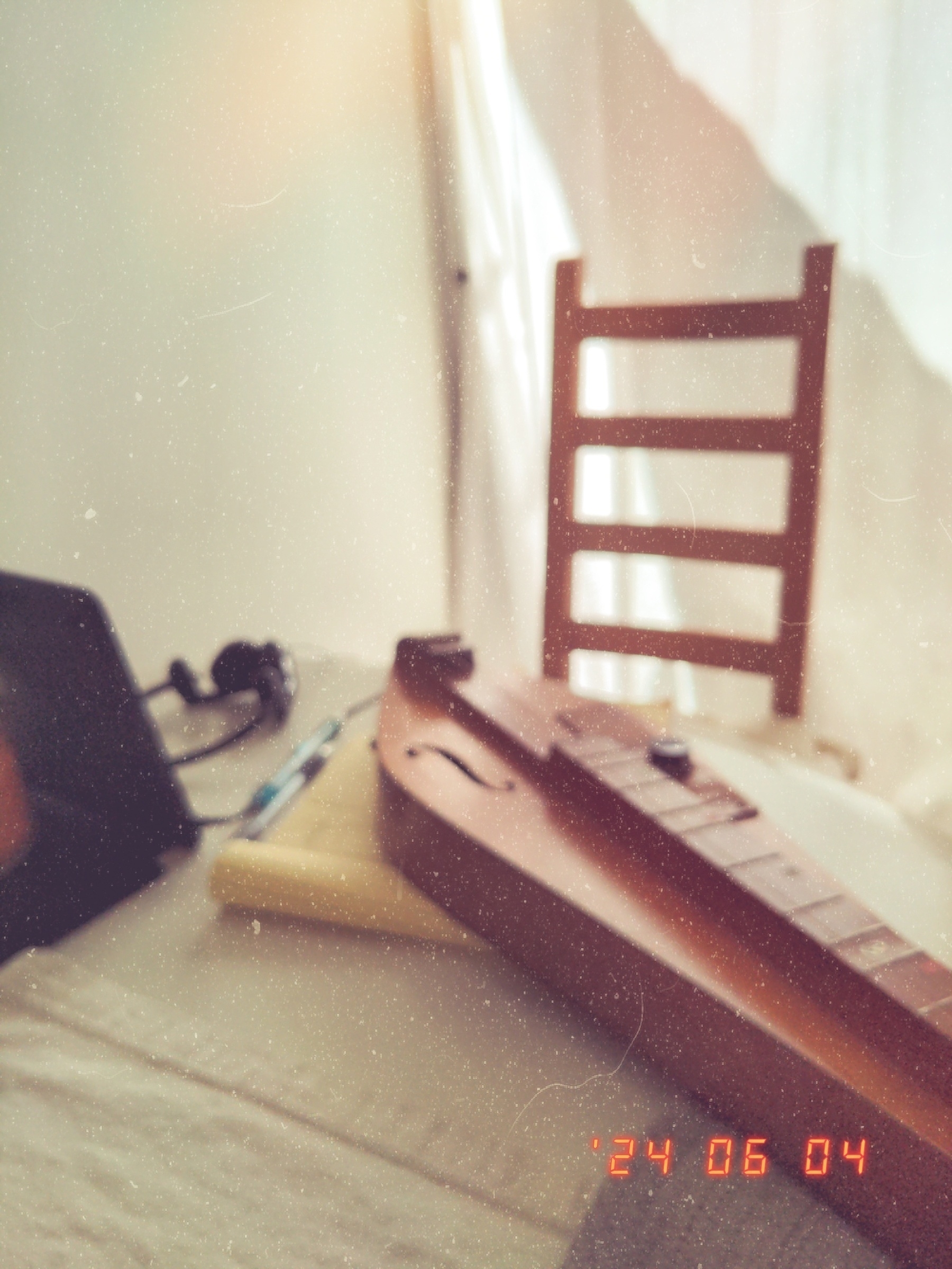 A mountain dulcimer sits on a round white table with some papers, headphones and a propped up iPad. A plain wooden chair is in the background and behind that a large window with sheer white curtains. Filtered sunlight is coming in through the window.