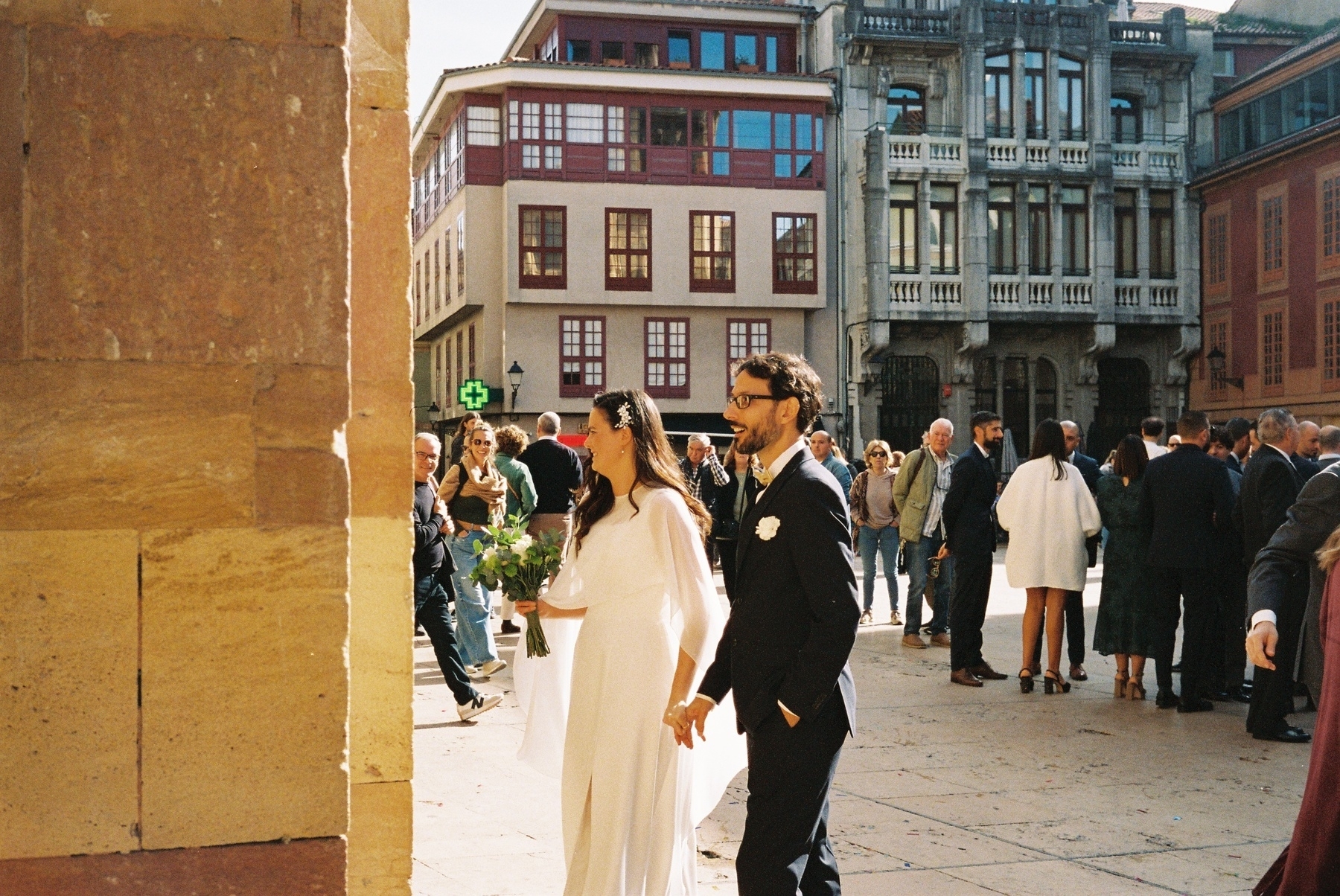 Auto-generated description: A couple in formal attire stands among a gathering of people outdoors near historic buildings.