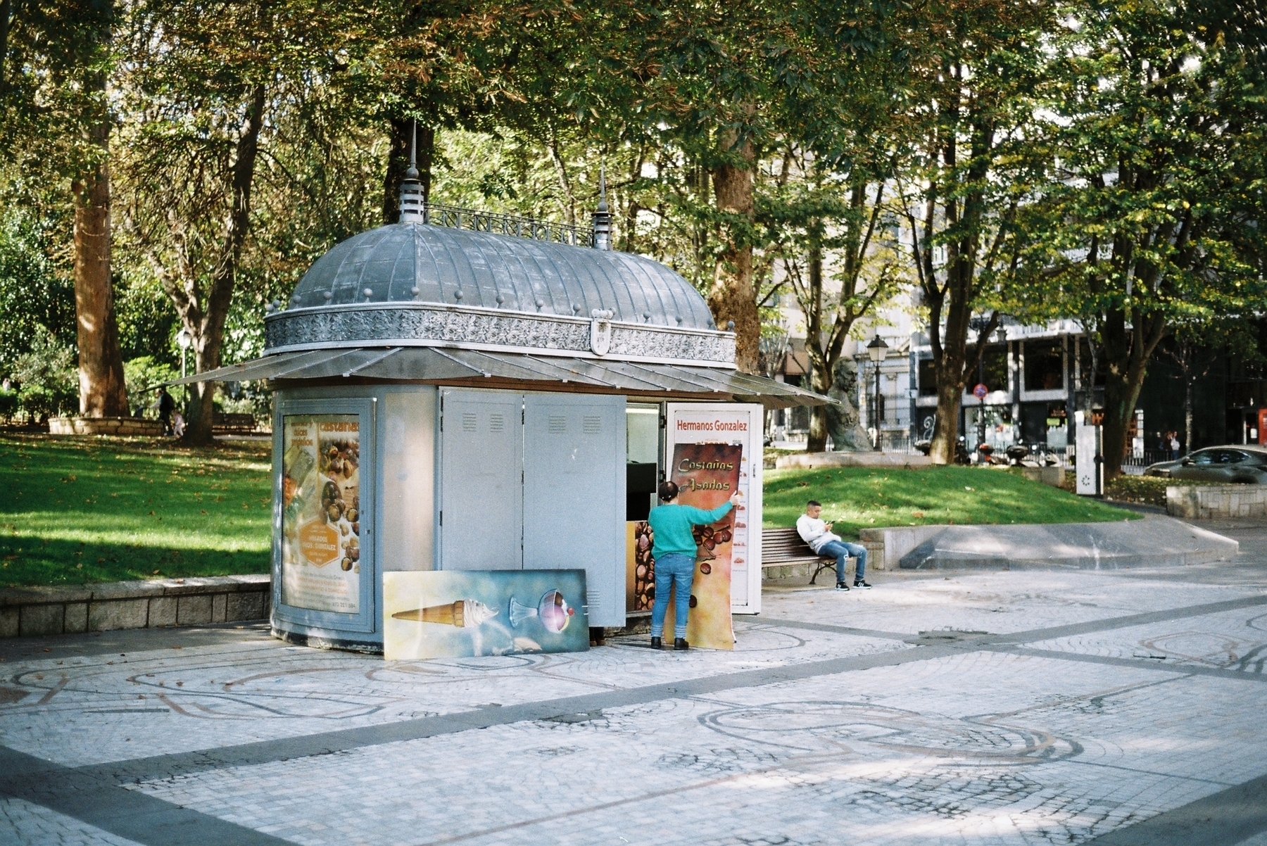 Auto-generated description: A person stands near a small kiosk in a park with trees and a person sitting on a bench nearby.