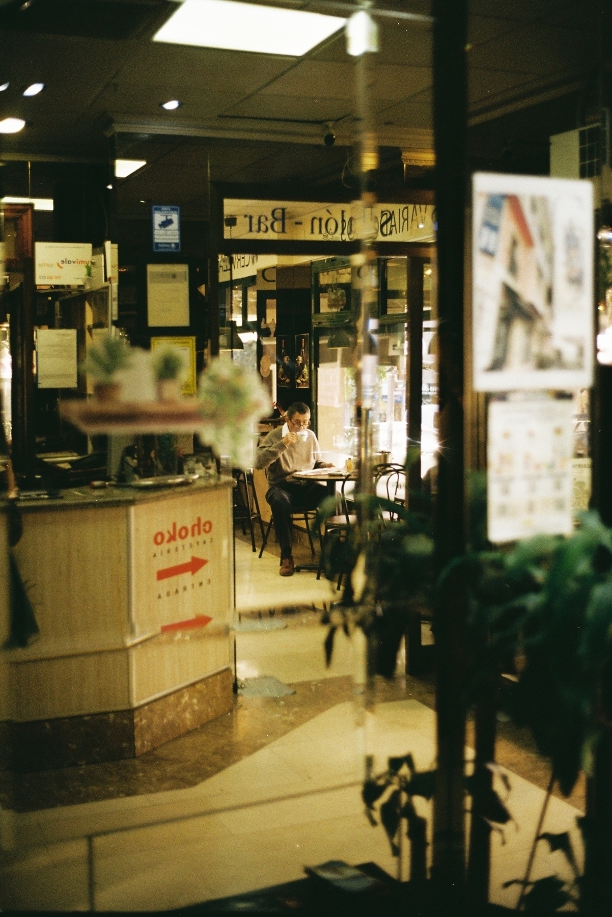 Auto-generated description: A person sitting alone at a table in a cozy café environment with reflections and various decorations surrounding them.