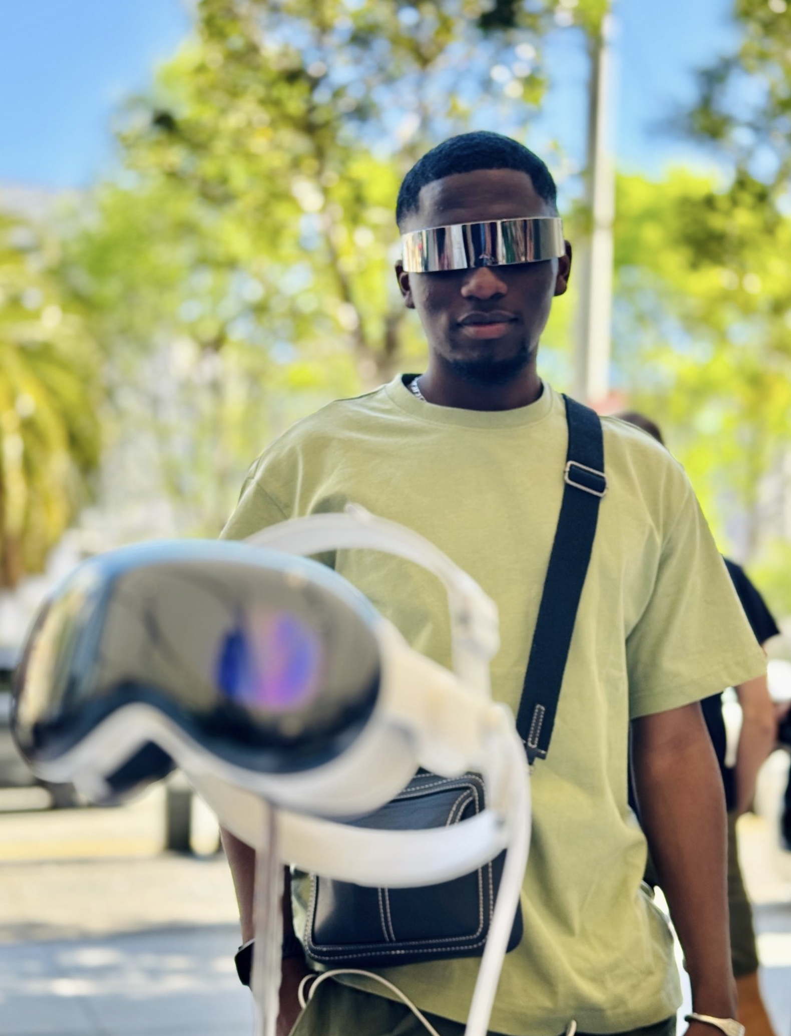 Auto-generated description: A person is wearing reflective futuristic glasses and standing outdoors, with blurred trees and a cityscape in the background.