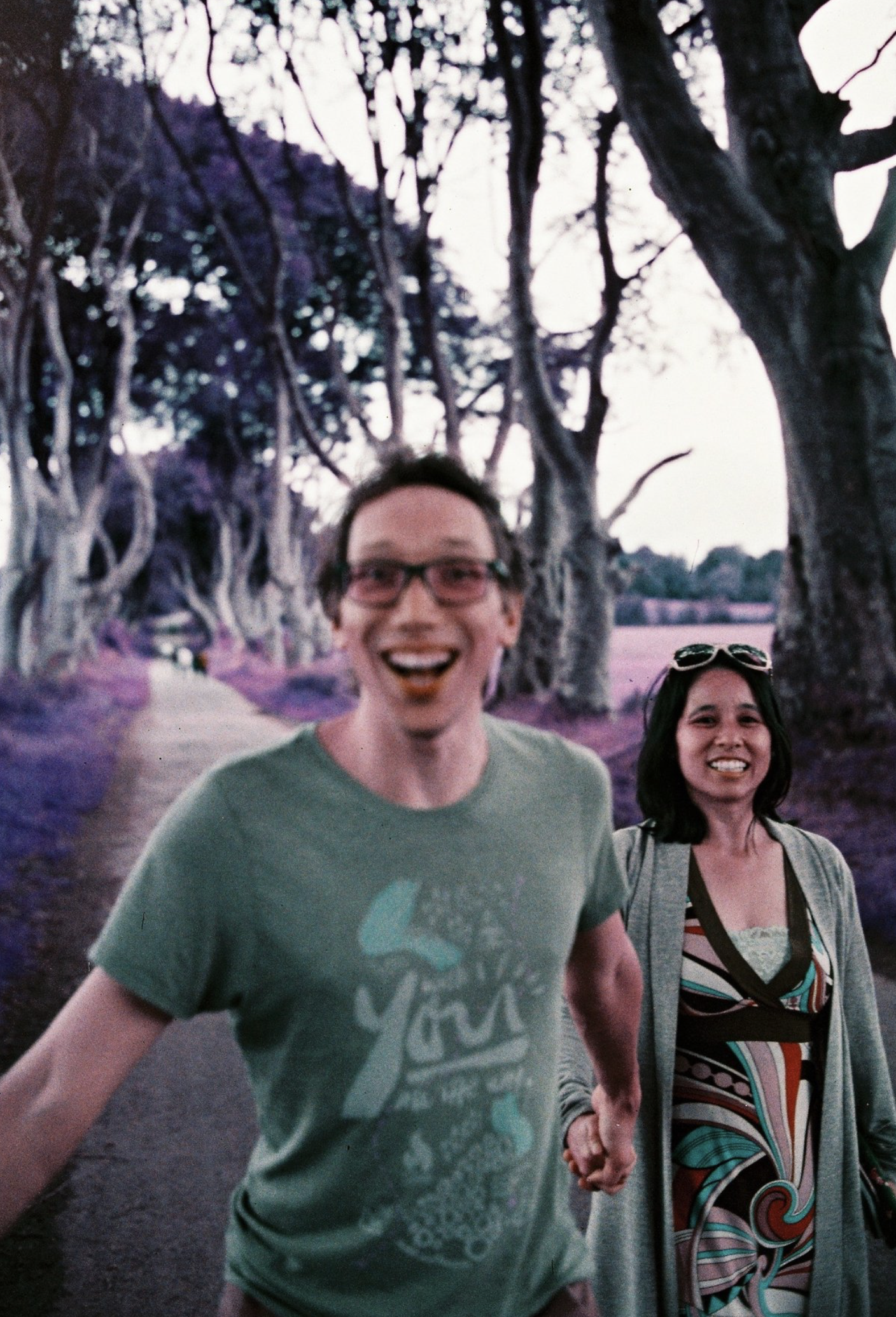 Auto-generated description: Two people are smiling and holding hands while walking down a tree-lined path with purple-tinted ground.