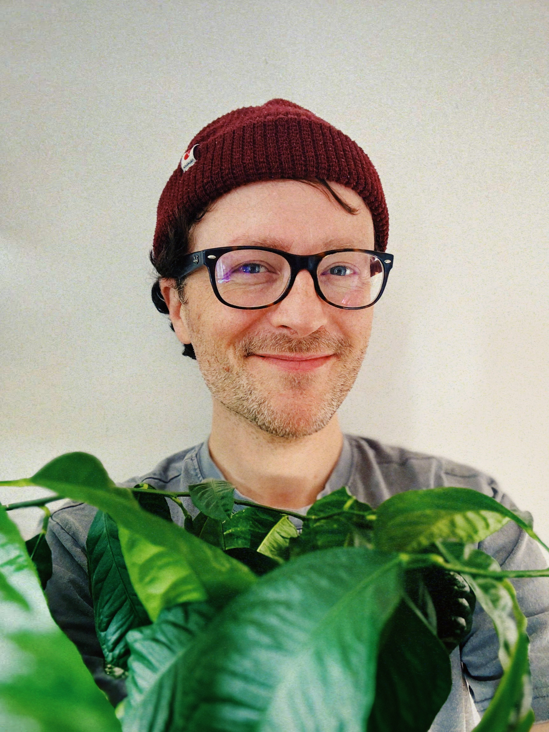 Auto-generated description: A person wearing glasses and a red beanie is smiling while standing behind a large green plant.