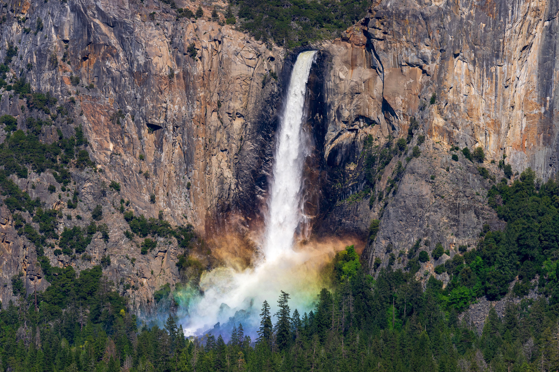 A waterfall pours over the lip of a cliff, crashing down onto rocks below, surrounded by trees. Where the water lands, a large spray of mist is illuminated by the setting sun, creating a rainbow effect throughout the mist.