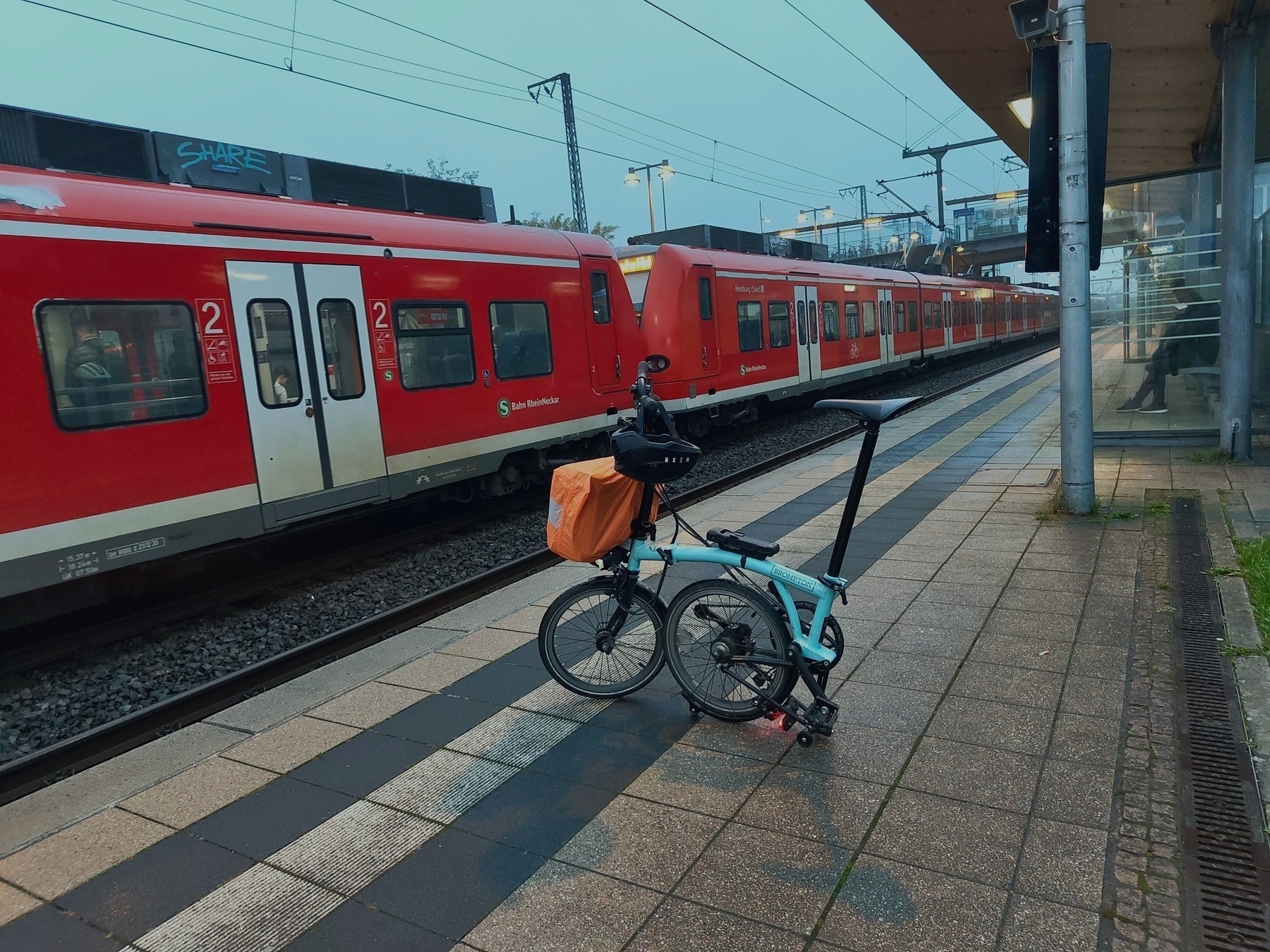 A half-folded Brompton bike sits on the platform across from a red S-Bahn, on a gloomy day