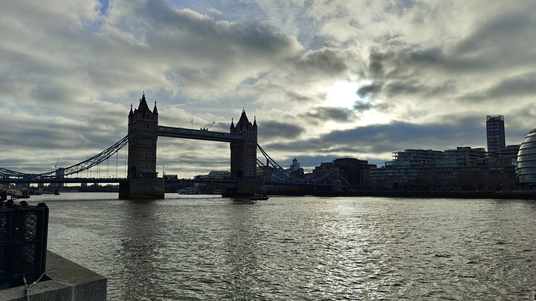 Auto-generated description: Tower Bridge stands against a cloudy sky over the Thames River, with the cityscape in the background.