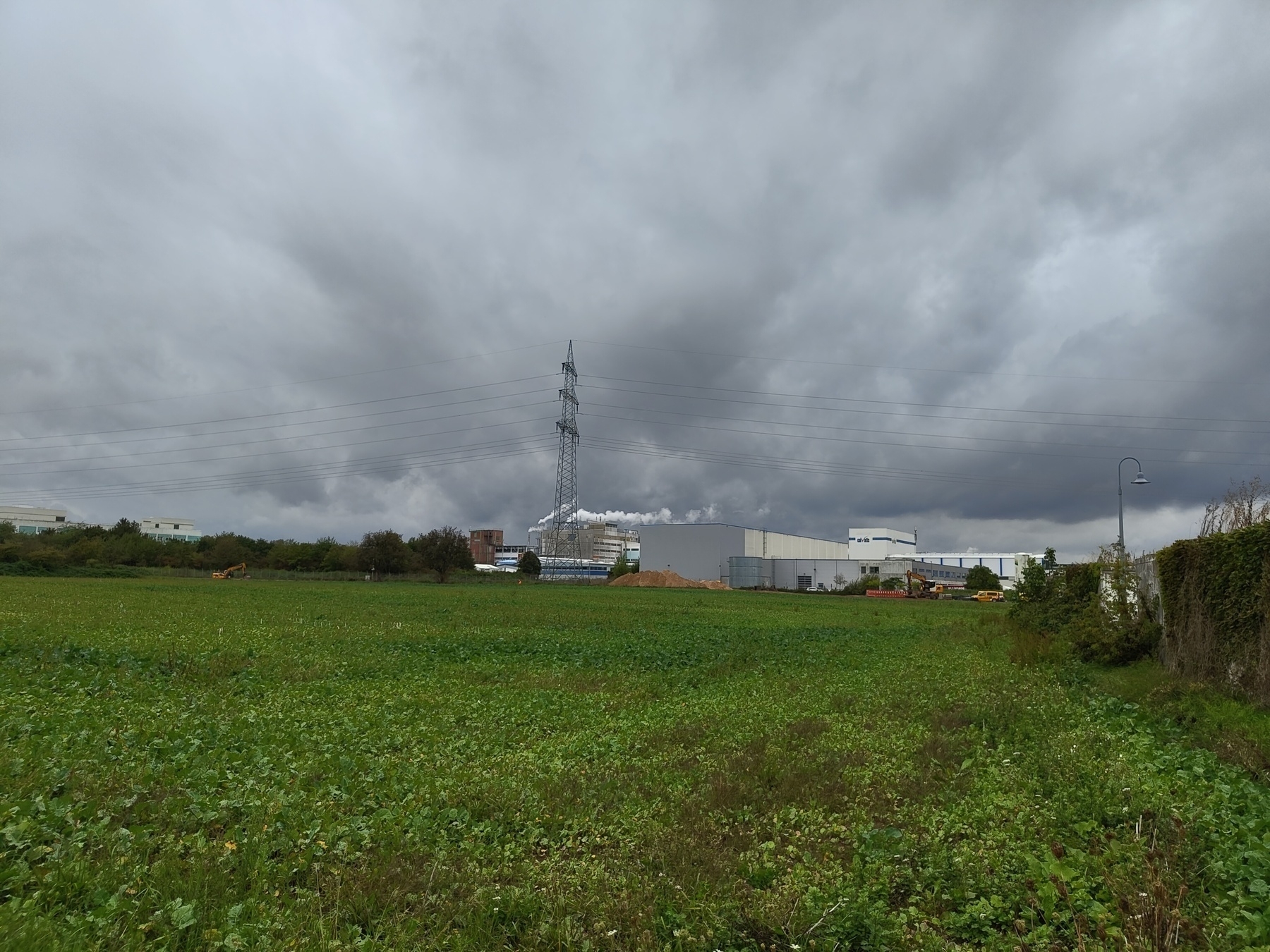 Auto-generated description: A stormy sky looms over a large green field with industrial buildings and a power line in the distance.