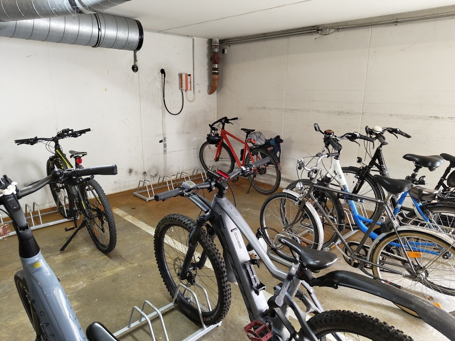 Auto-generated description: Several bicycles are parked in an indoor storage area with a cement floor and exposed ventilation ducting.