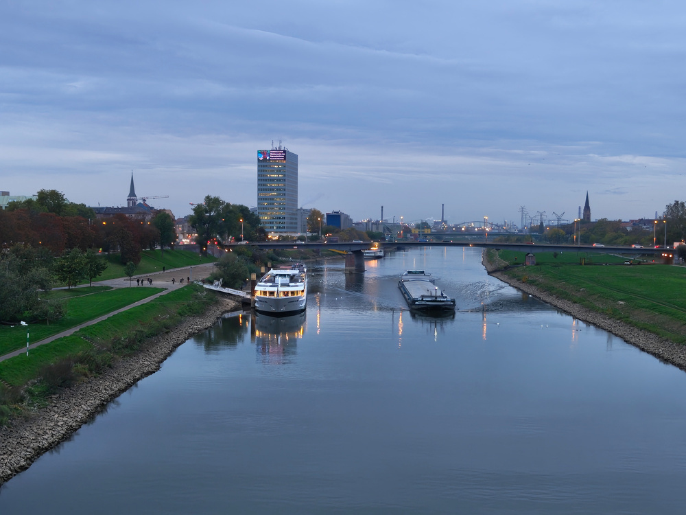A river scene at dusk features two boats, a riverside path, and a cityscape with a prominent tower in the background.