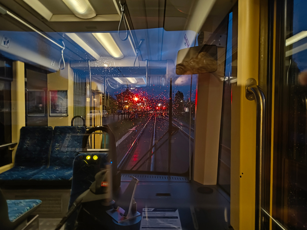 A view from inside a tram shows raindrops on the window and red lights outside during evening, with a reflection of the seats and interior.