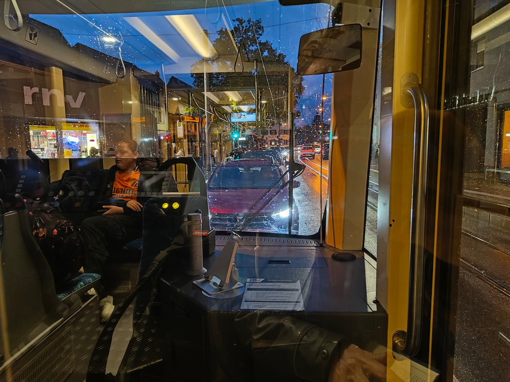 Inside a tram, a passenger sits opposite the driver's cabin as city lights and traffic are visible through the wet window at night.