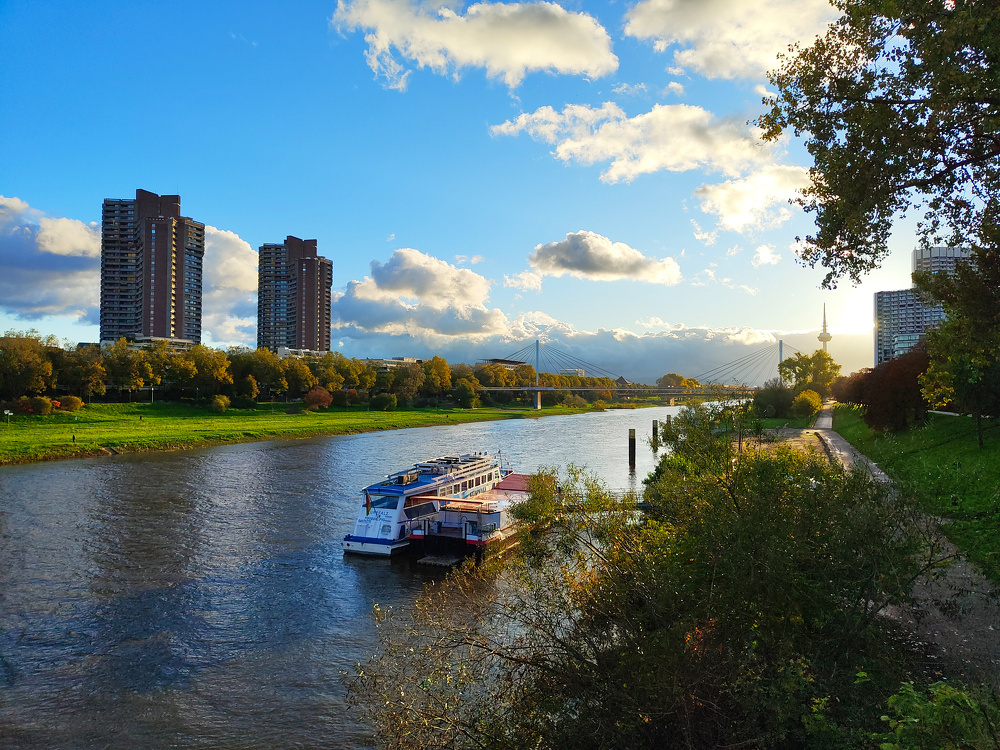 A serene riverside scene features a docked boat, modern buildings, lush greenery, and a partly cloudy sky.