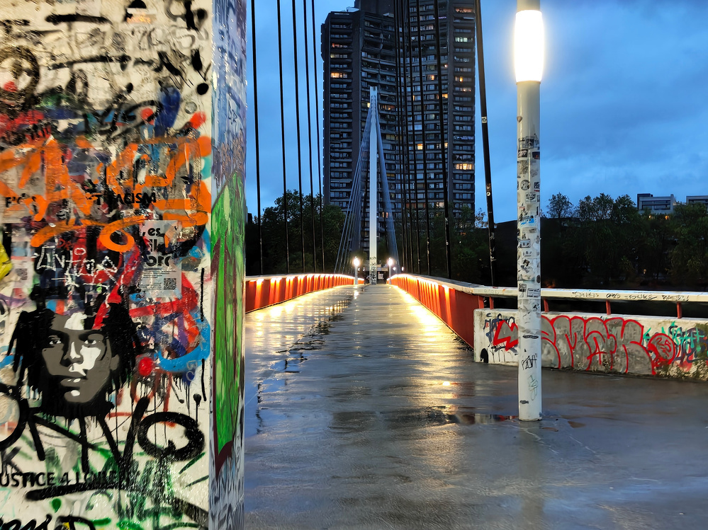 A rain-soaked bridge adorned with vibrant graffiti leads towards a tall building under a cloudy evening sky.